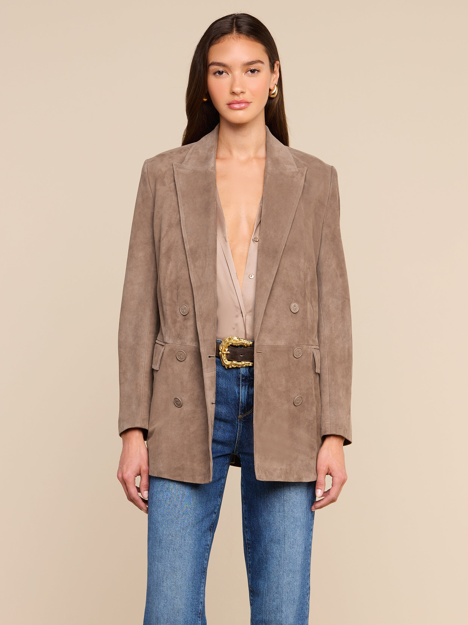A woman wearing the Aimee Oversized Suede Blazer in taupe over a beige blouse, paired with blue jeans and a textured belt, stands against a plain light background, looking forward with her hands at her sides.
