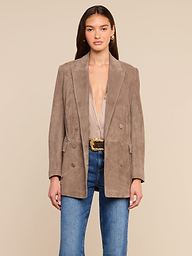 A woman wearing the Aimee Oversized Suede Blazer in taupe over a beige blouse, paired with blue jeans and a textured belt, stands against a plain light background, looking forward with her hands at her sides.