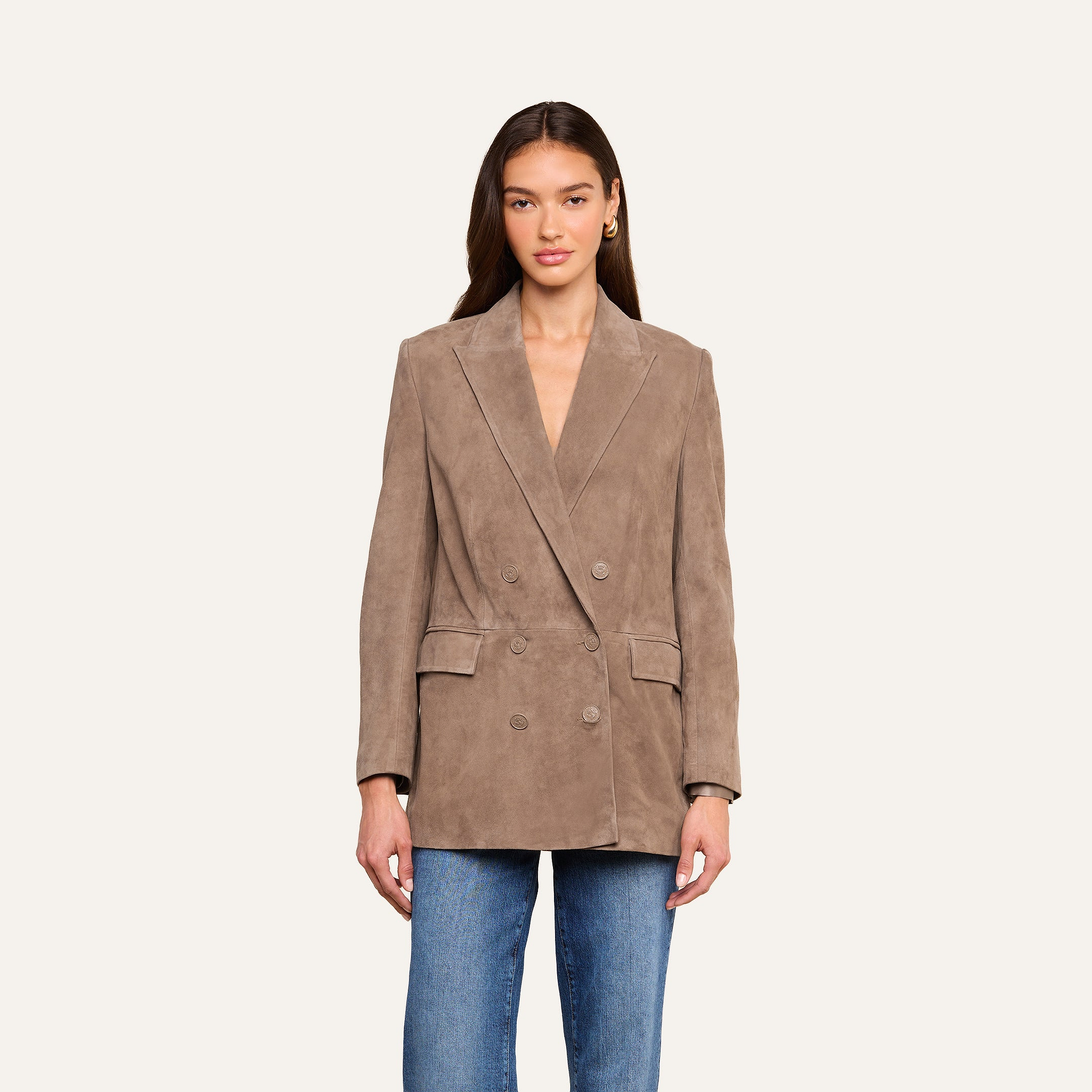 A woman wearing the Aimee Oversized Suede Blazer in tan suede, featuring wide lapels and an oversized fit, pairs it with blue jeans as she stands against a plain light background, looking straight ahead with her arms relaxed by her sides.