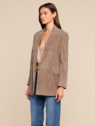 A woman wearing the Aimee Oversized Suede Blazer in tan over a beige top and blue jeans stands against a plain light background, looking slightly to the side.