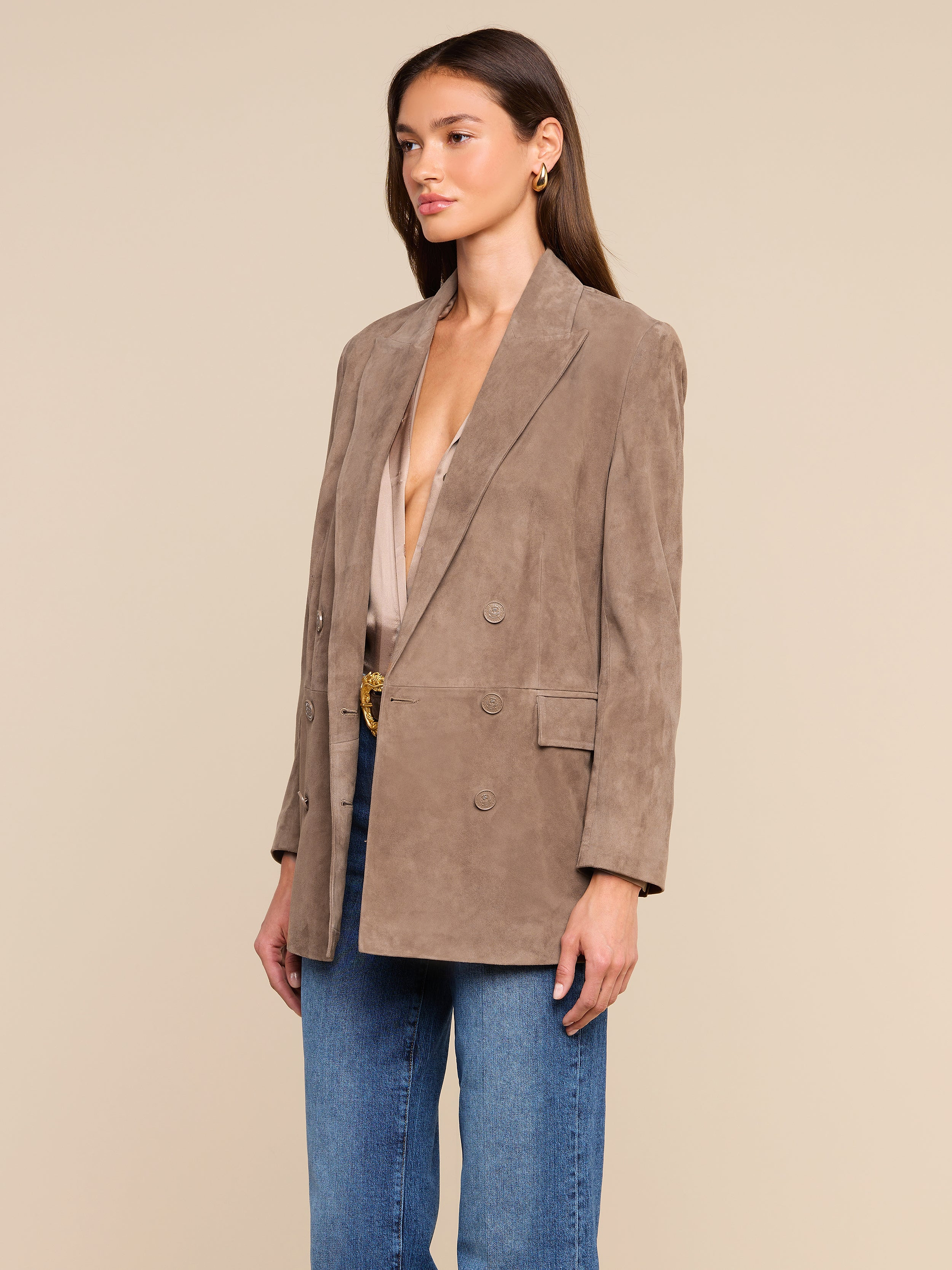 A woman wearing the Aimee Oversized Suede Blazer in tan over a beige top and blue jeans stands against a plain light background, looking slightly to the side.