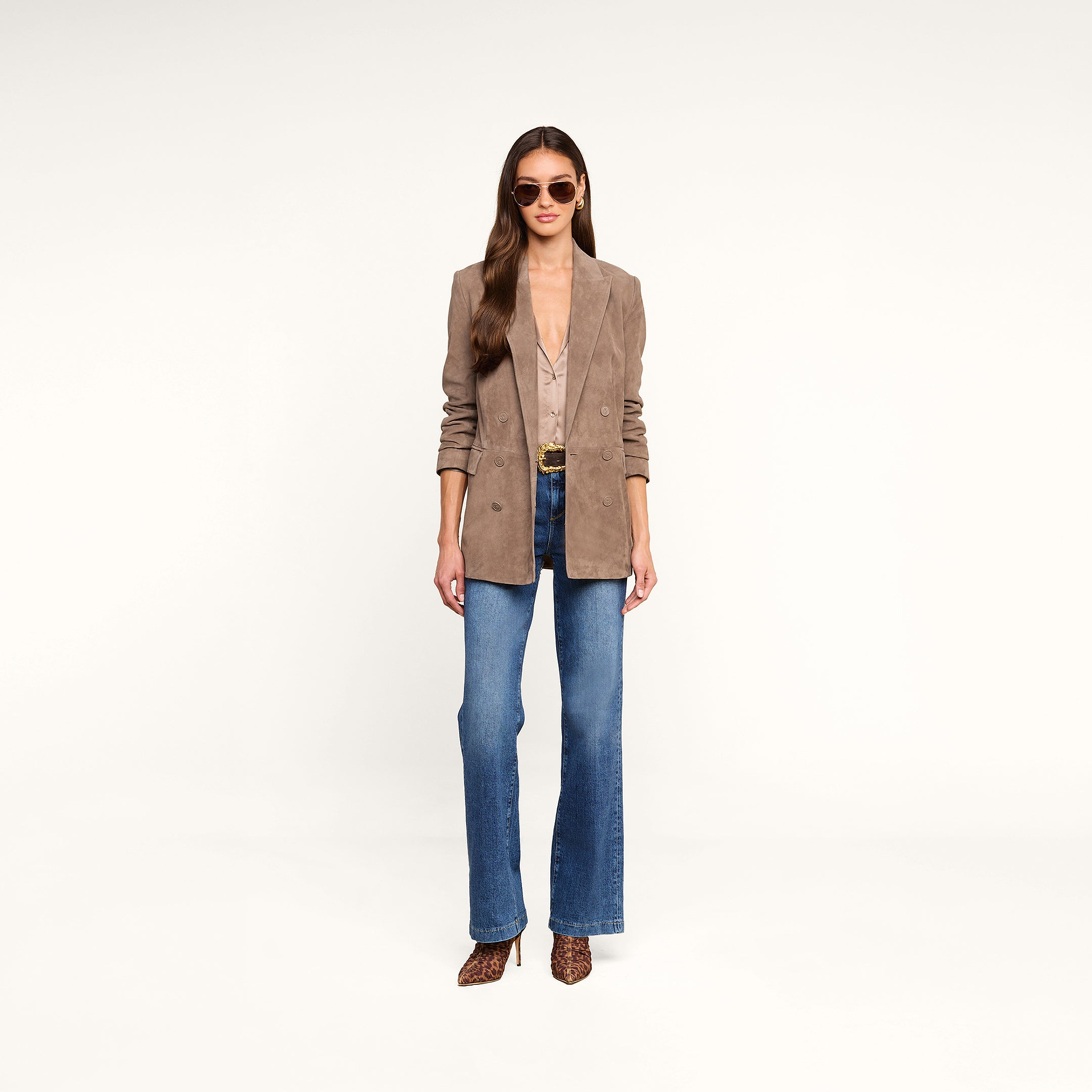 A woman stands facing forward against a plain white background, wearing sunglasses, the Aimee Oversized Suede Blazer in tan over a deep V-neck blouse, blue flared jeans, and leopard print shoes.
