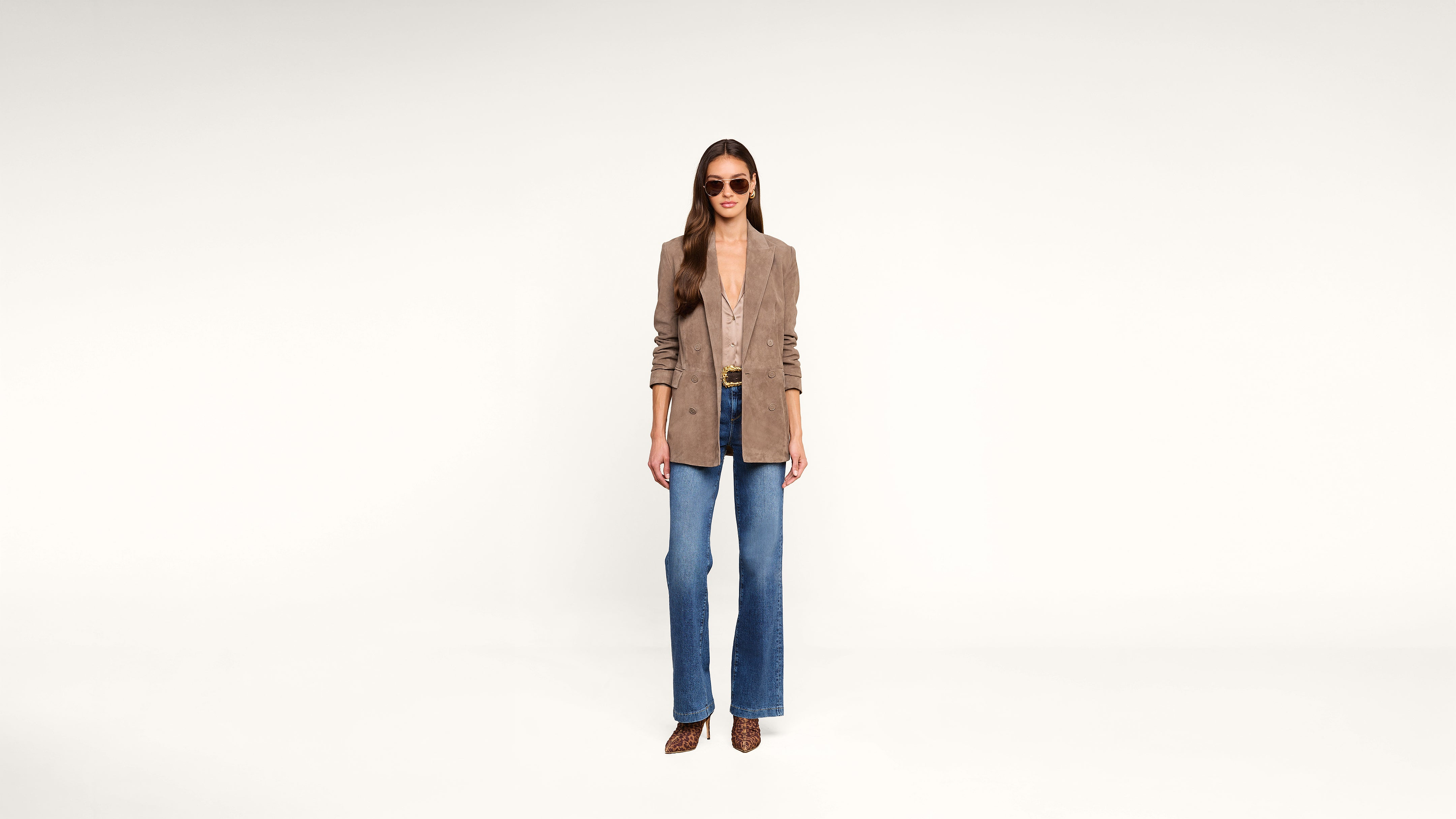 A woman stands against a white background wearing sunglasses, a tan blazer, a low-cut top, blue jeans, and brown heels.
