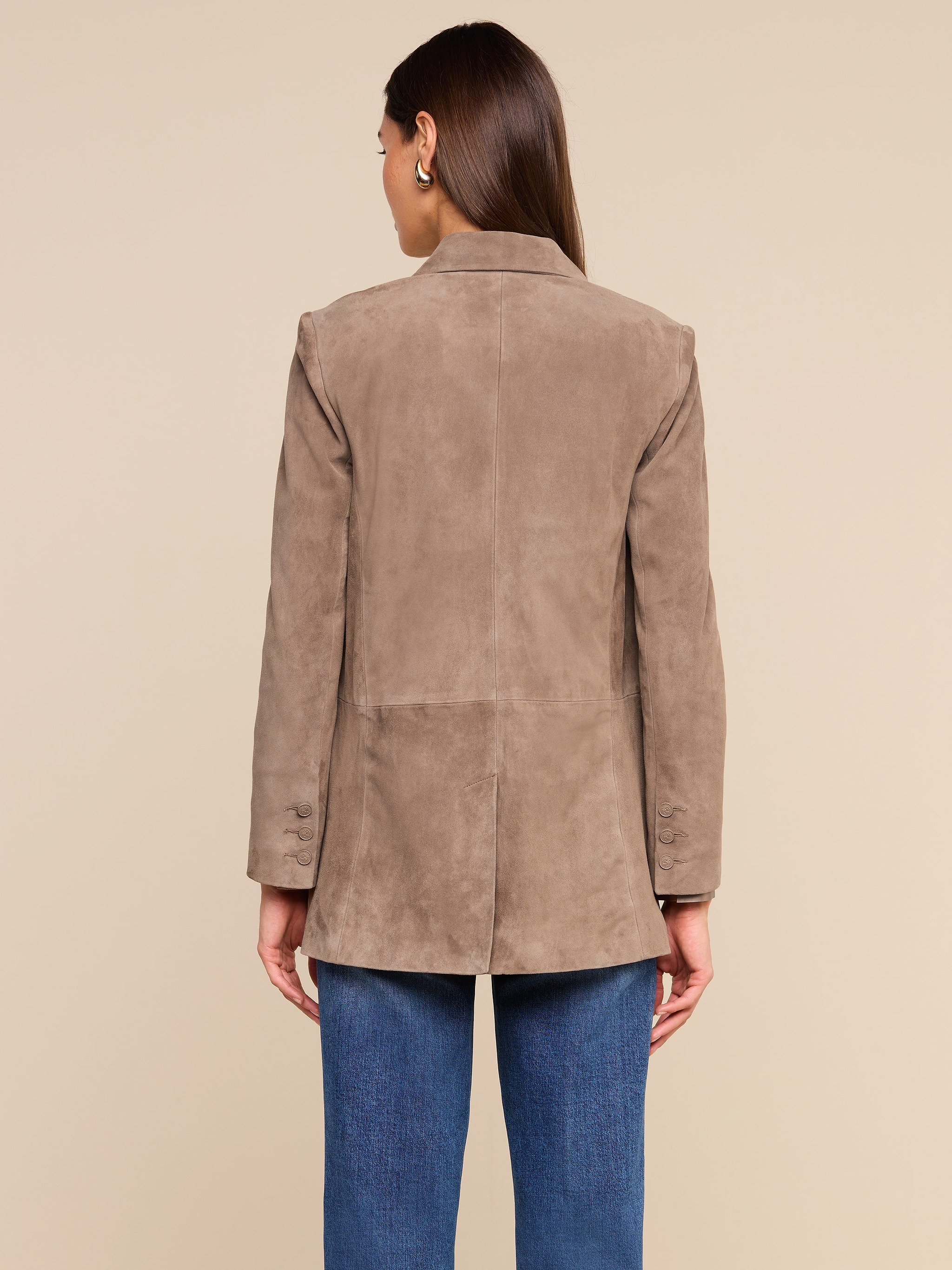 A woman stands with her back to the camera against a plain, light background, wearing the Aimee Oversized Suede Blazer in tan with blue jeans. Her straight brown hair falls to her shoulders.
