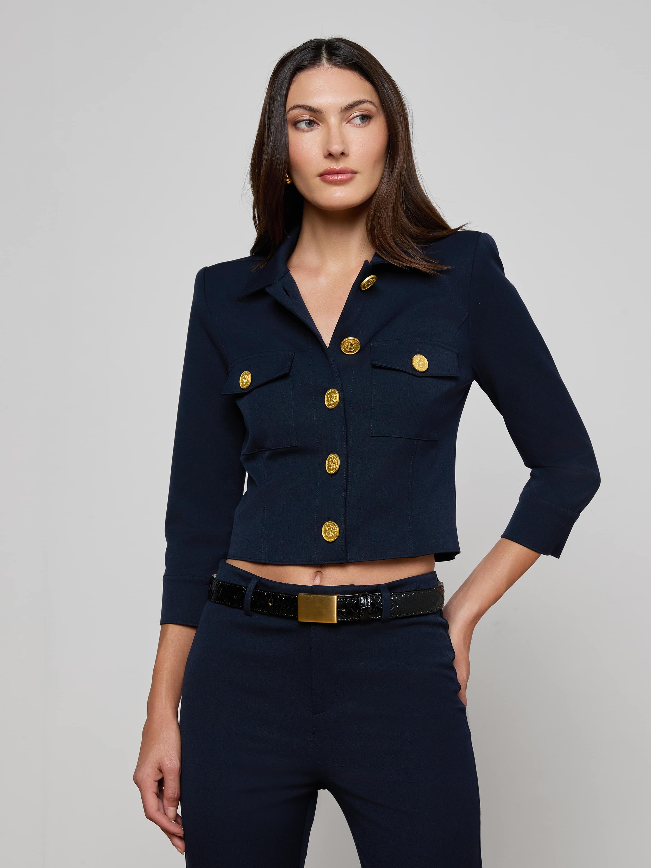 A woman wearing the Kumi Cropped Jacket in midnight navy, featuring gold buttons and three-quarter sleeves, stands against a plain light background. The jacket, crafted from stretch suit fabric for comfort and style, is paired with matching navy pants as she looks slightly to her left with her left hand resting near her pocket.