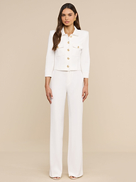 A woman stands against a plain white background, wearing the Kumi Cropped Jacket crafted from stretch suiting with three-quarter sleeves and gold-embossed dome buttons, paired with matching wide-leg white pants.