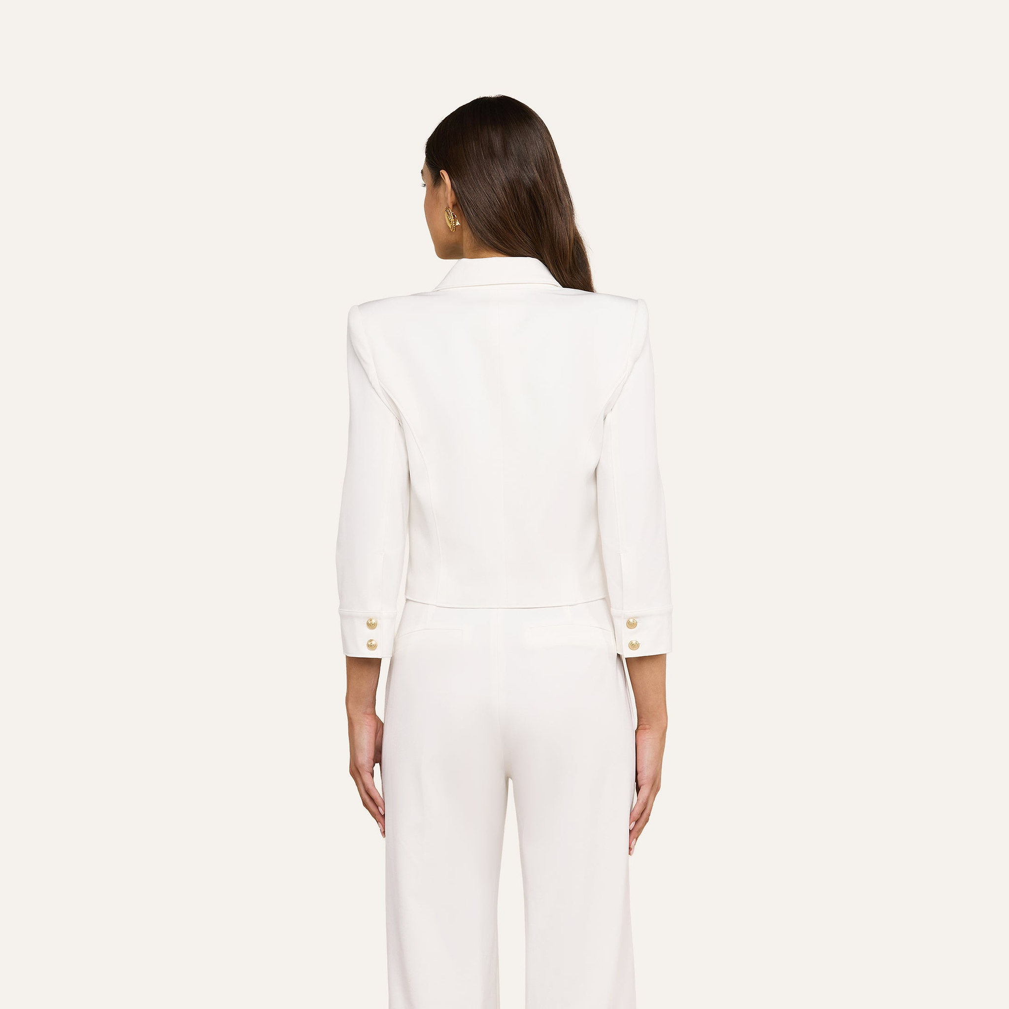 A woman with long brown hair stands facing away, wearing the Kumi Cropped Jacket in white stretch-suiting fabrication with gold embossed dome buttons and matching white pants against a plain light background.