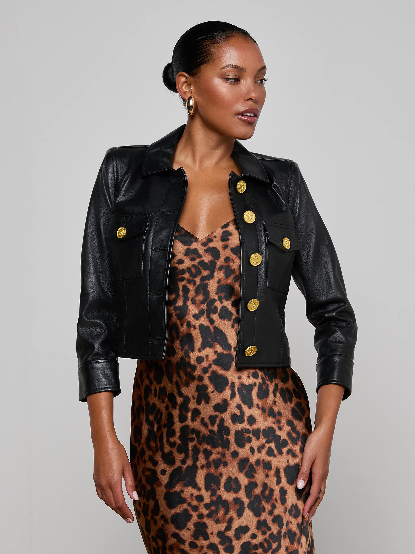 A woman poses against a plain light background, wearing the Kumi Cropped Leather Jacket with gold buttons over a sleeveless leopard print dress. The three-quarter sleeve leather jacket adds an edgy touch to her chic ensemble.