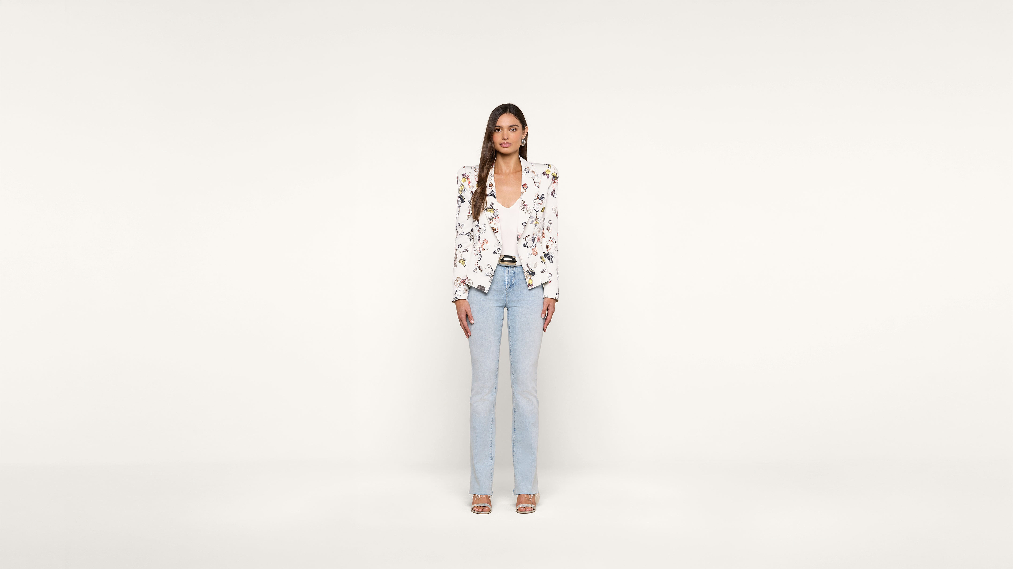 Woman stands facing forward, wearing a white floral blazer, white top, light blue jeans, and open-toed heels, against a plain white background.
