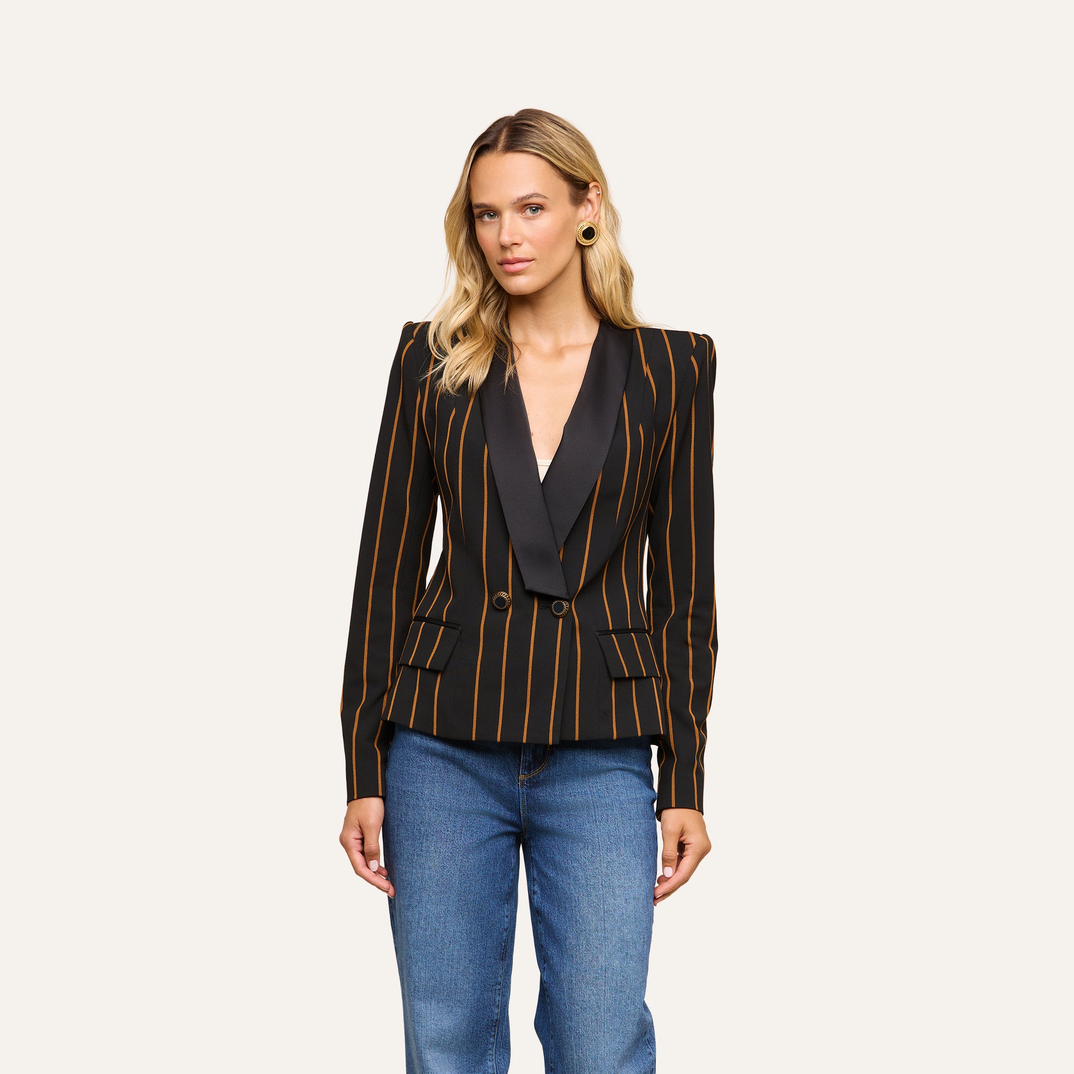A woman with long blonde hair wears the Imogen Satin Lapel Blazer—a black double-breasted blazer featuring gold vertical stripes and dark satin lapels—paired with blue jeans, standing against a plain white background.