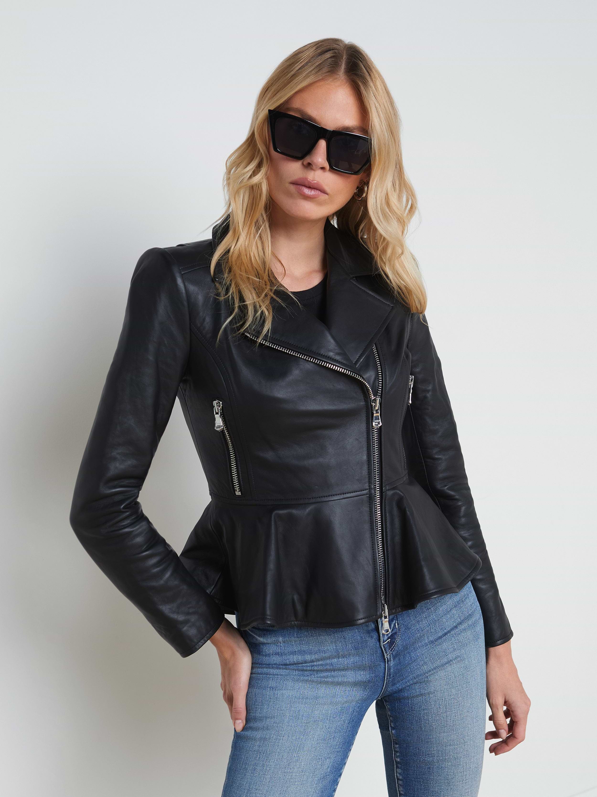 A woman wears the Lyric Leather Peplum Jacket—crafted in black lambskin leather with zip details—paired with blue jeans and dark sunglasses, as she stands against a plain light background.