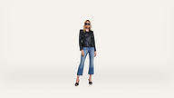A woman wearing sunglasses, a black leather jacket, blue cropped jeans, and black heels stands confidently against a plain white background.
