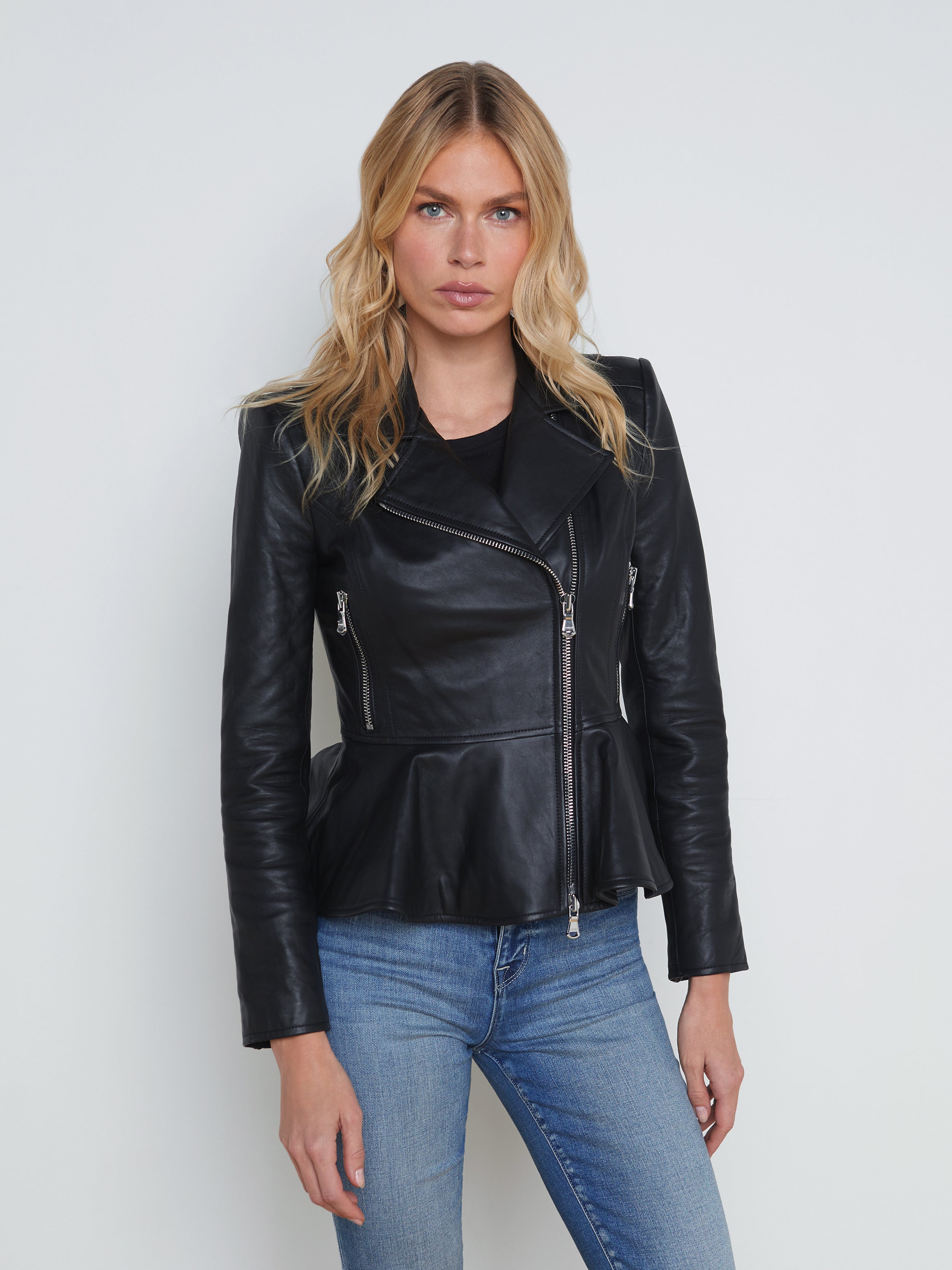 A woman with wavy blonde hair wears the Lyric Leather Peplum Jacket in black lambskin leather and blue jeans, standing against a plain off-white background, facing forward with a neutral expression.