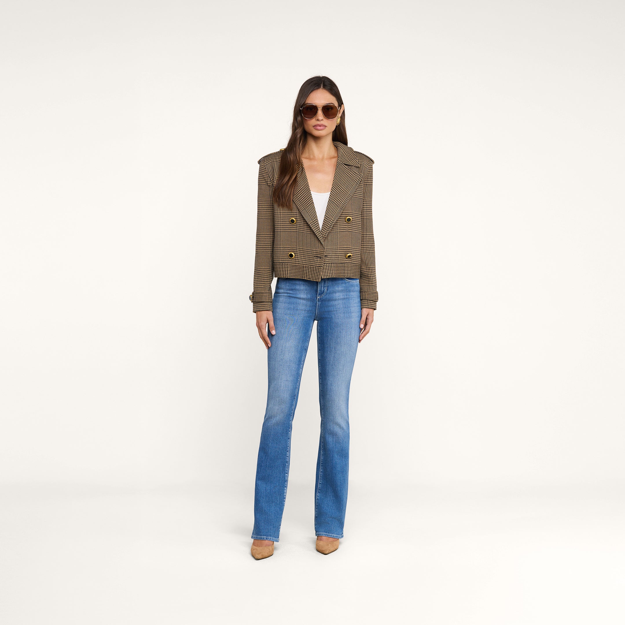 A woman wearing sunglasses, the Xoey Cropped Trench Coat styled as a double-breasted jacket, a white top, blue jeans, and tan heels stands against a plain white background.
