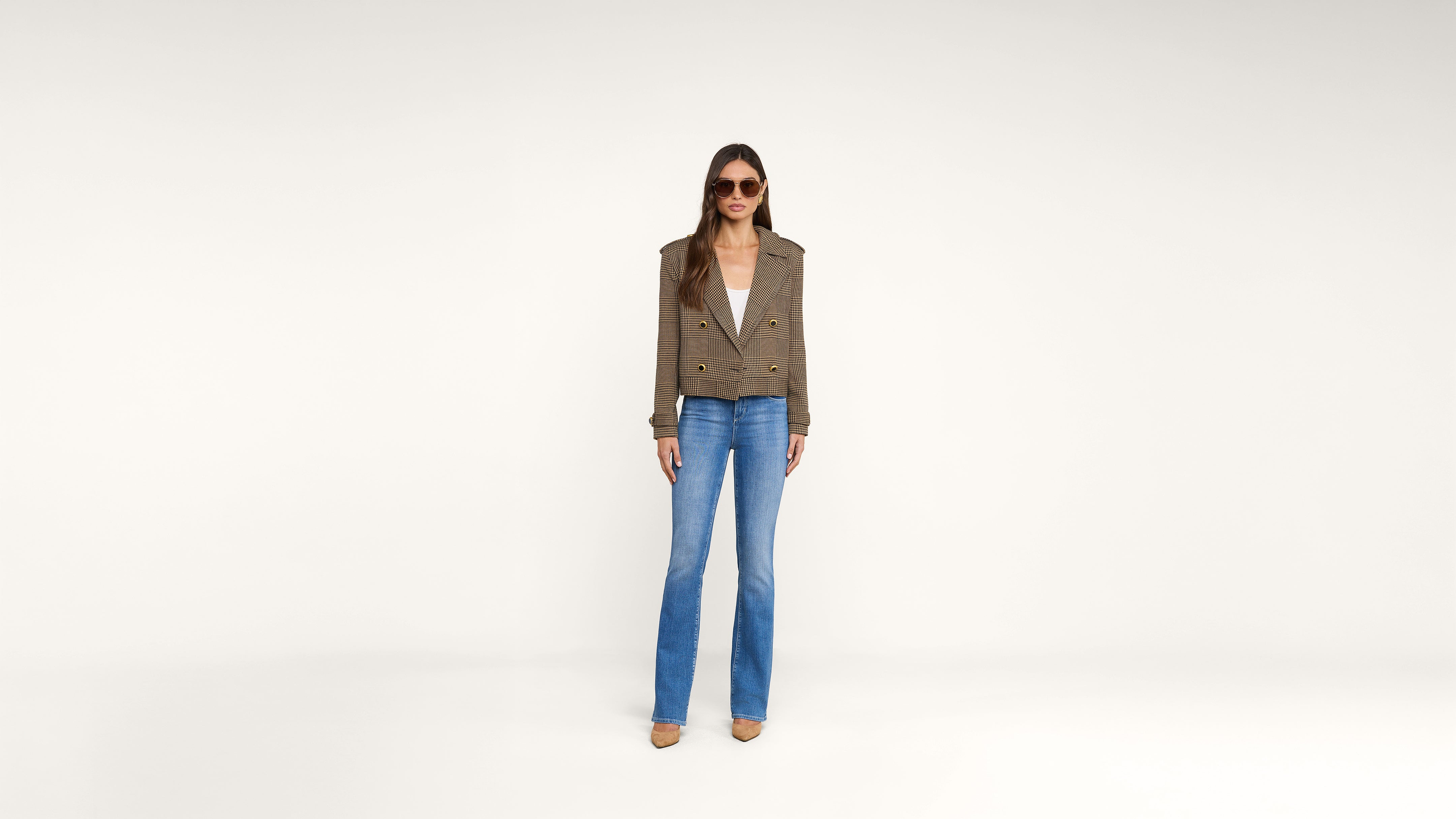 A woman wearing sunglasses, a brown plaid blazer, white top, blue jeans, and brown shoes stands against a plain white background.