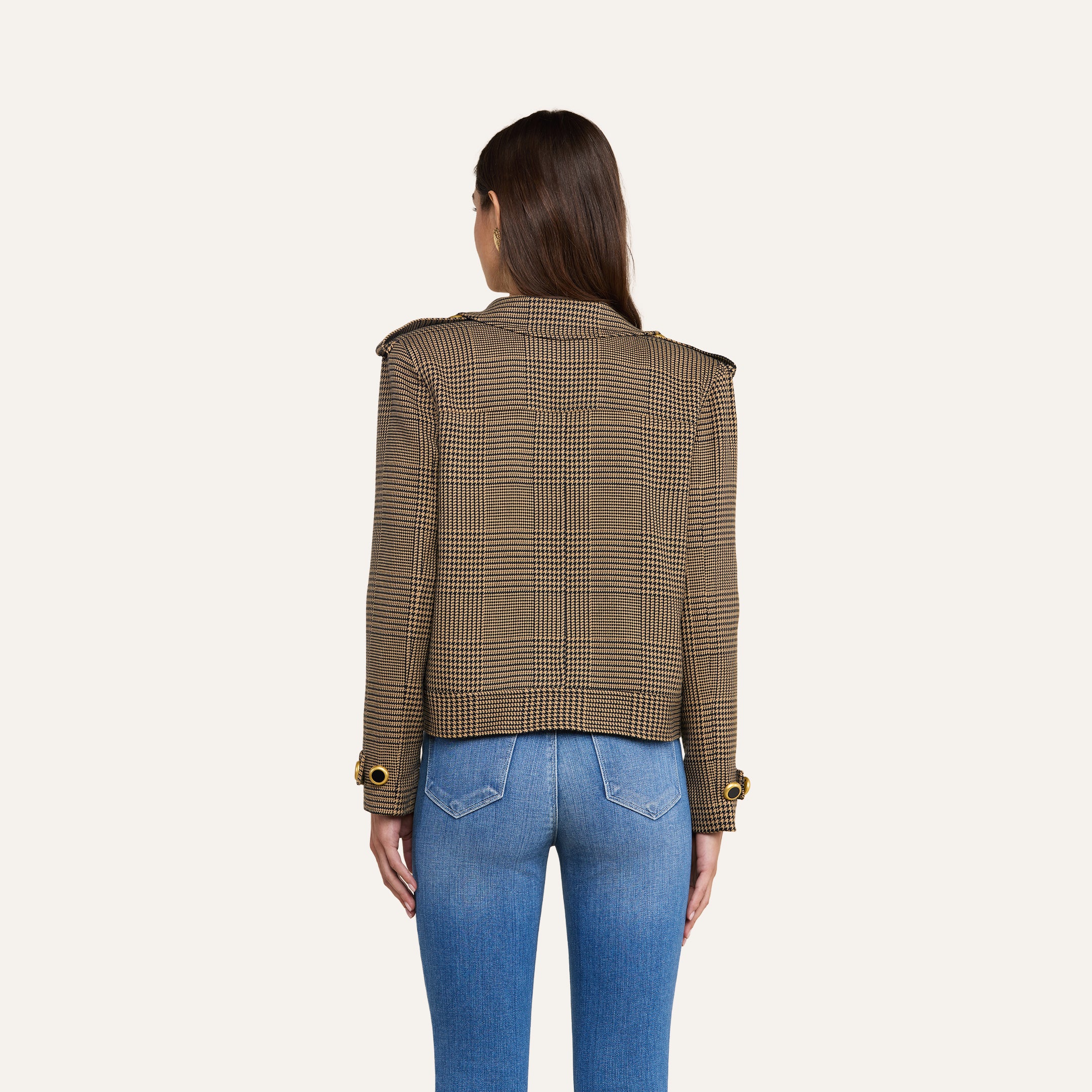 A woman stands with her back to the camera, wearing the Xoey Cropped Trench Coat—a brown plaid double-breasted jacket with gold buttons—paired with light blue fitted jeans, against a plain light background.