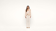 A woman stands facing forward against a plain white background, wearing a light beige jacket, matching pants, and tan shoes.