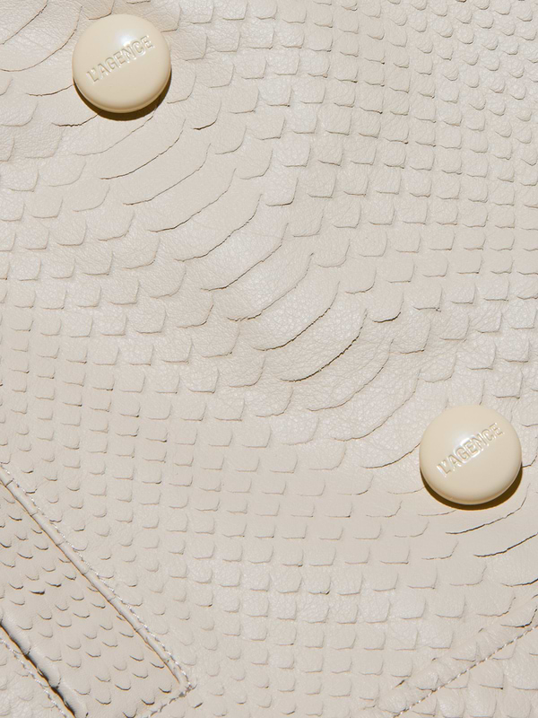 Close-up of the cream-colored, snake-embossed leather surface of the Xoey Cropped Leather Trench Coat, featuring two round beige buttons labeled HABENICE.