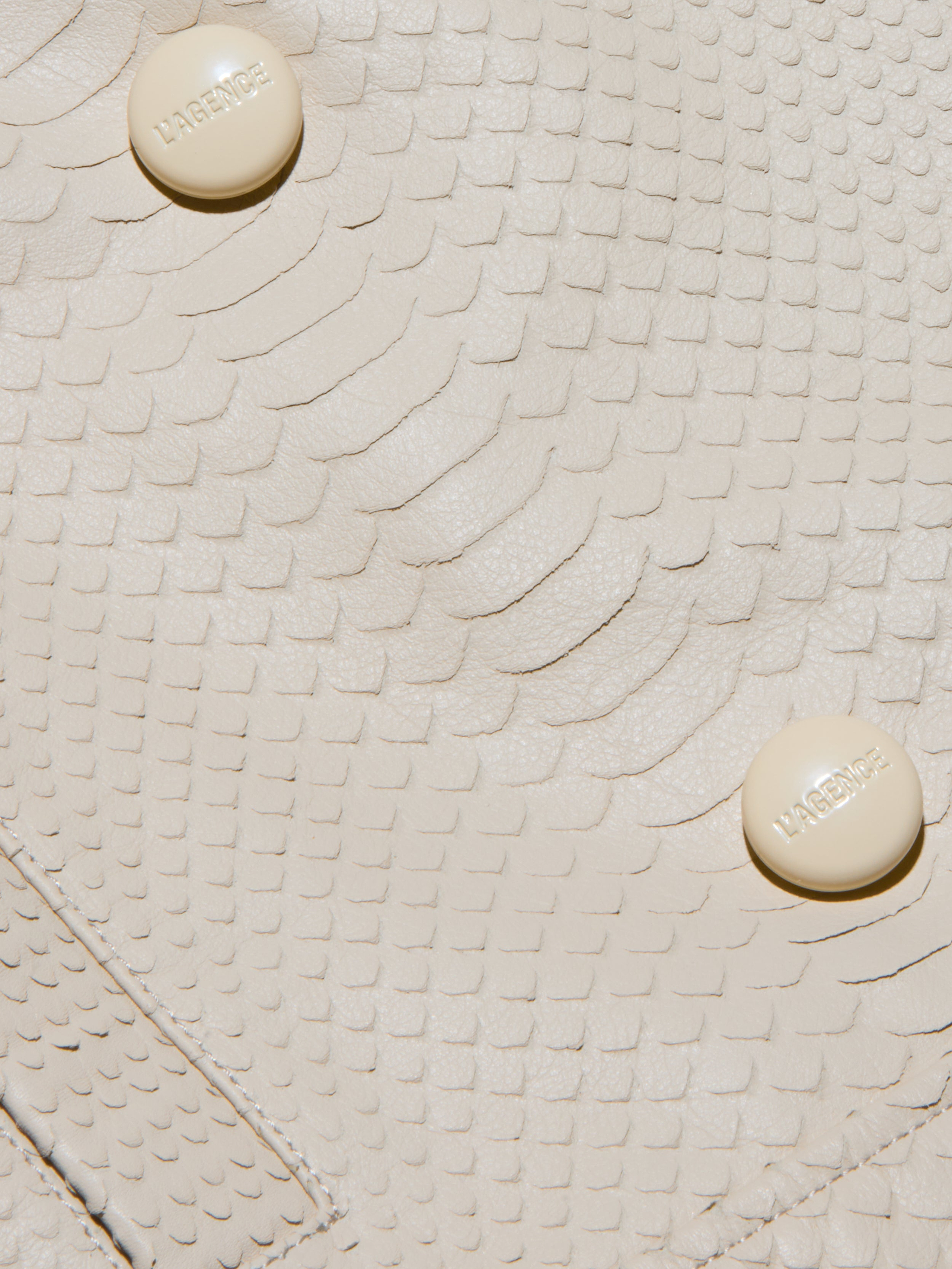 Close-up of the cream-colored, snake-embossed leather surface of the Xoey Cropped Leather Trench Coat, featuring two round beige buttons labeled HABENICE.
