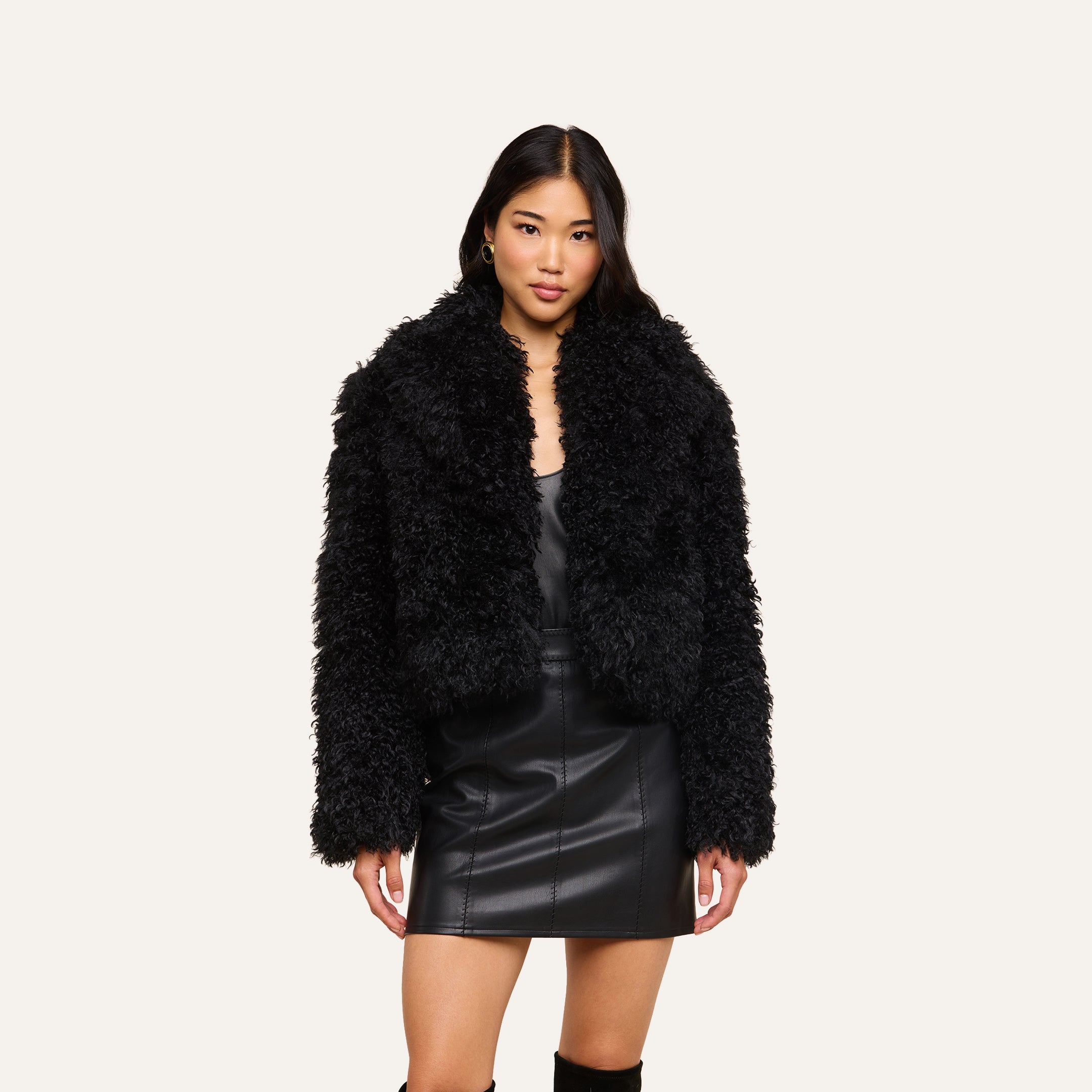 A woman stands facing forward against a plain light background, wearing the Aura Cropped Faux Fur Jacket over a black leather dress and knee-high black boots. She has long dark hair, a neutral expression, and poses confidently.