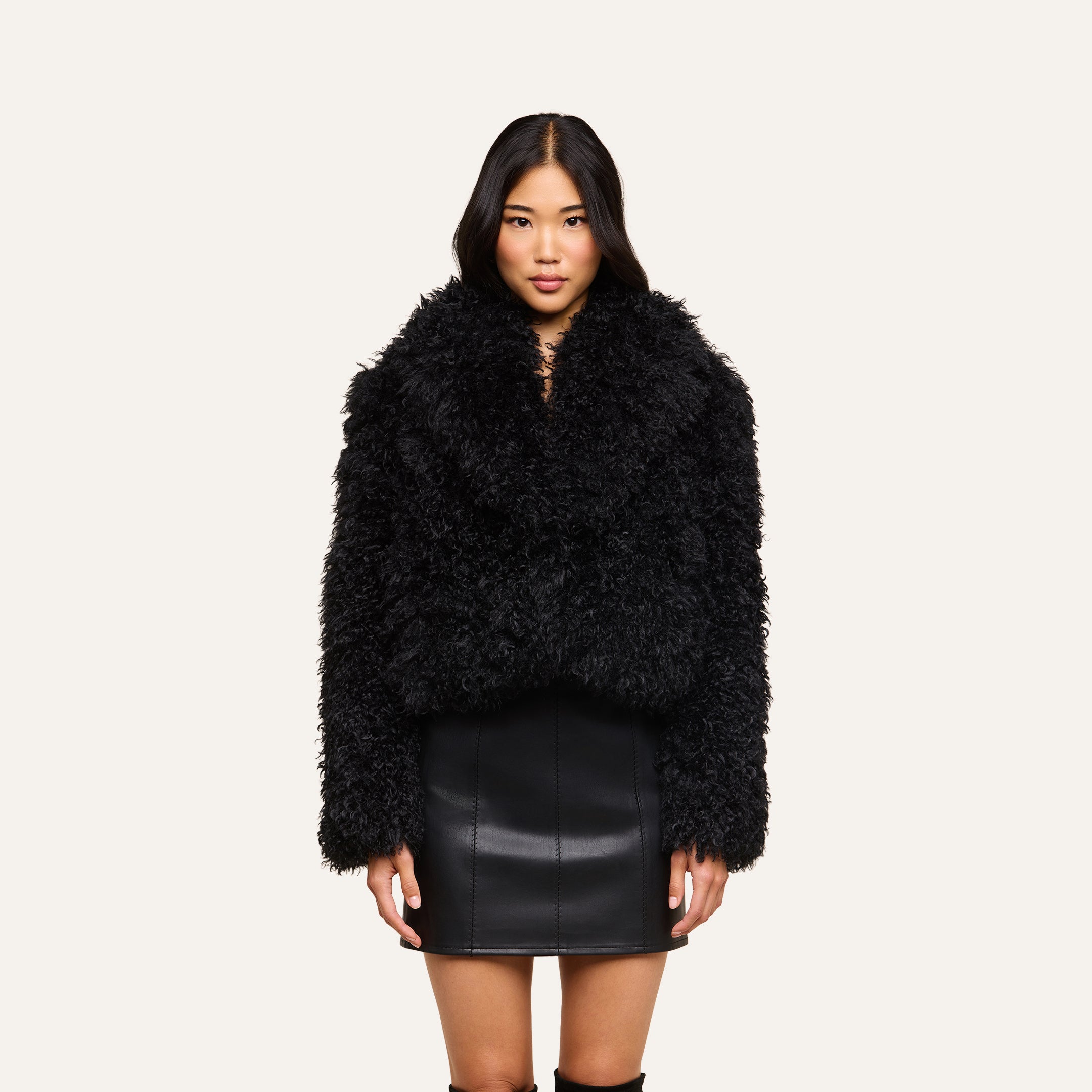 A woman wearing the Aura Cropped Faux Fur Jacket and a black leather skirt stands facing forward against a plain light background.