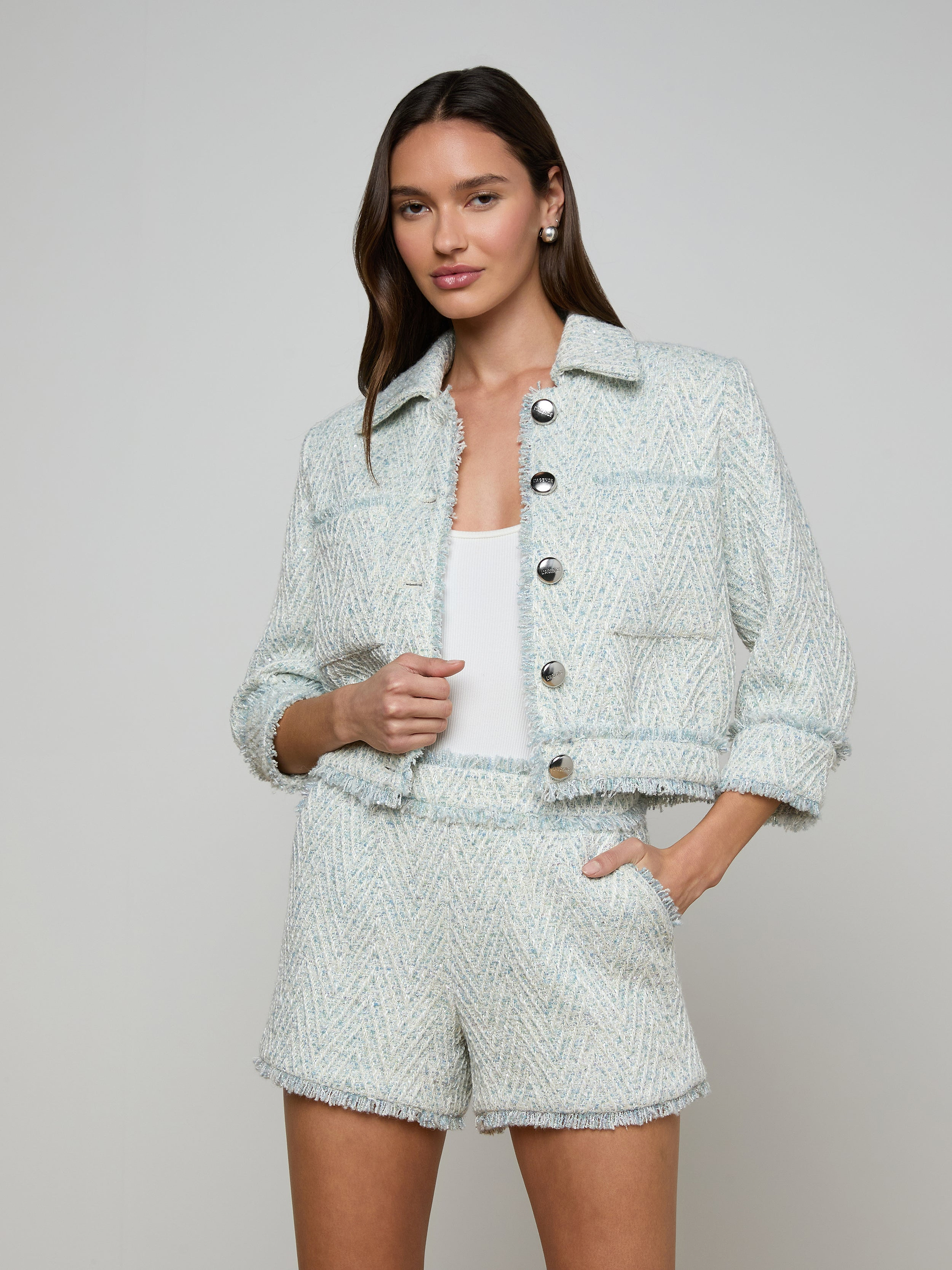 A woman wears the Jia Fringed Tweed Jacket in light blue chevron with matching shorts over a white top, standing with one hand in her pocket and the other holding the jacket, against a plain light-colored background.