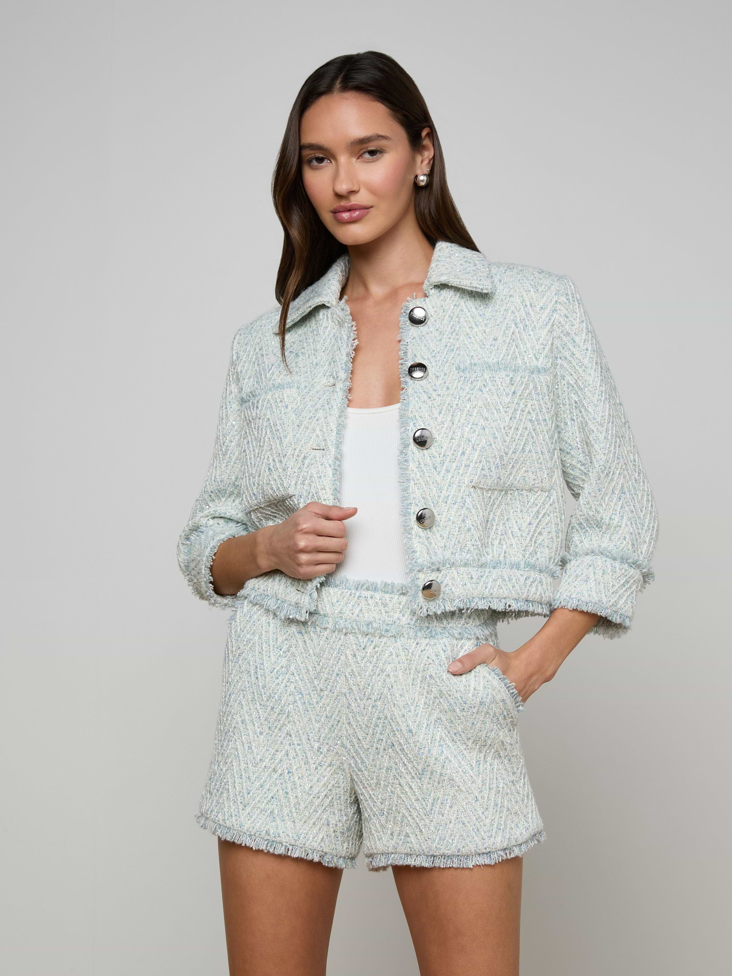 A woman wears the Jia Fringed Tweed Jacket in light blue chevron with matching shorts over a white top, standing with one hand in her pocket and the other holding the jacket, against a plain light-colored background.