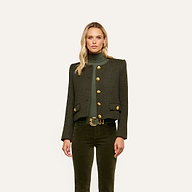A woman stands against a plain background, wearing an olive-green turtleneck and the Zell Tweed Jacket—a collarless tweed jacket with gold buttons—paired with matching pants featuring a gold-buckled belt.