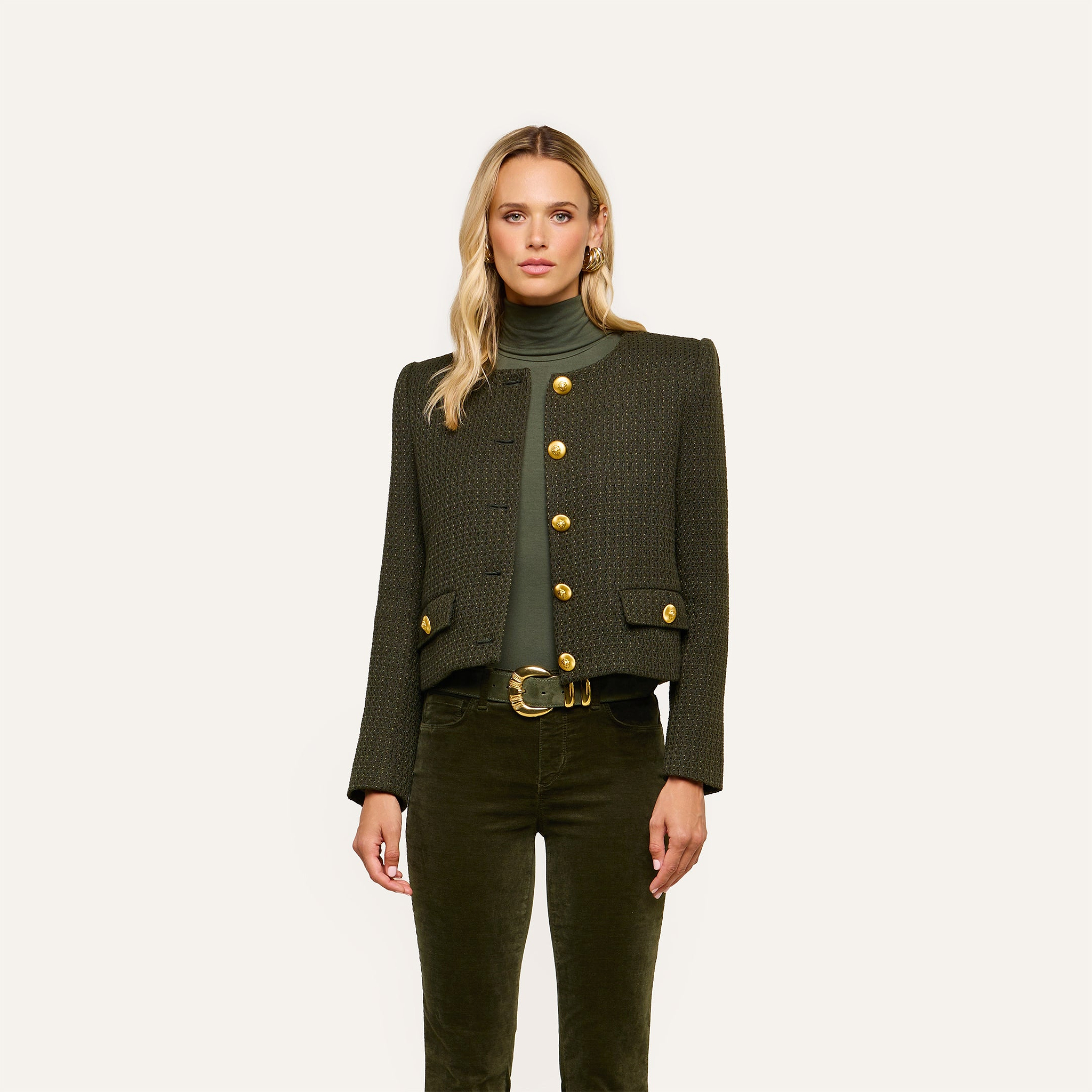 A woman stands against a plain background, wearing an olive-green turtleneck and the Zell Tweed Jacket—a collarless tweed jacket with gold buttons—paired with matching pants featuring a gold-buckled belt.