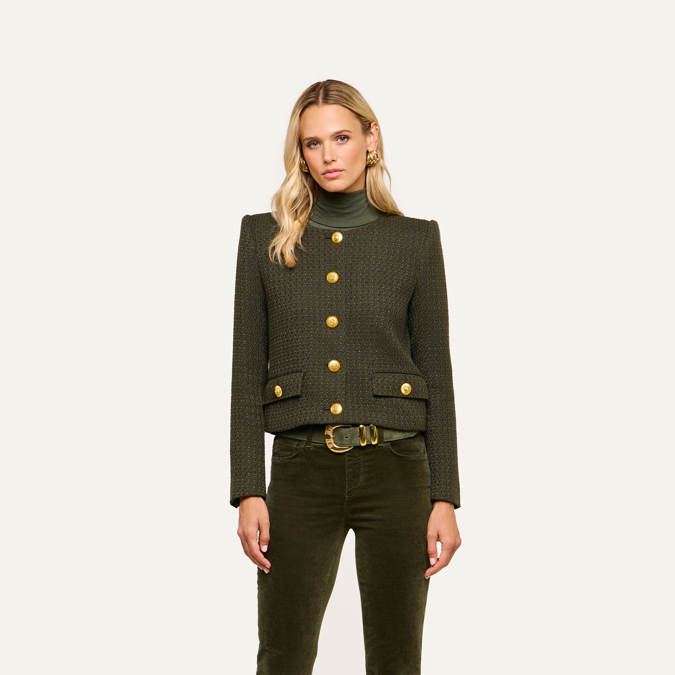 A woman with long blonde hair wears the Zell Tweed Jacket—a collarless dark green piece with gold buttons—paired with green pants and a belt, standing against a plain light background.