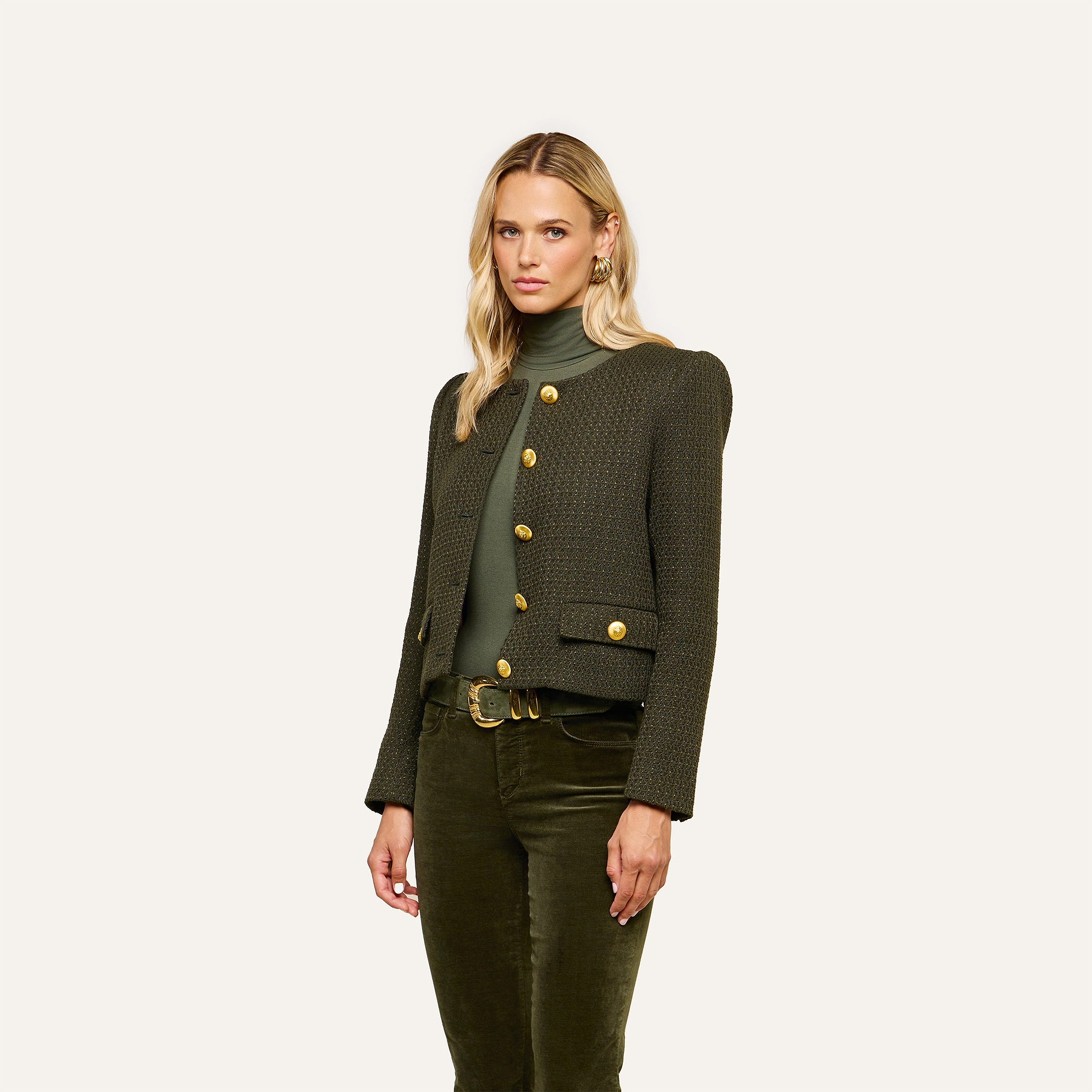A woman with long blonde hair wears the Zell Tweed Jacket—a dark green style with gold buttons—paired with a matching green turtleneck and green velvet pants, standing against a plain light background.