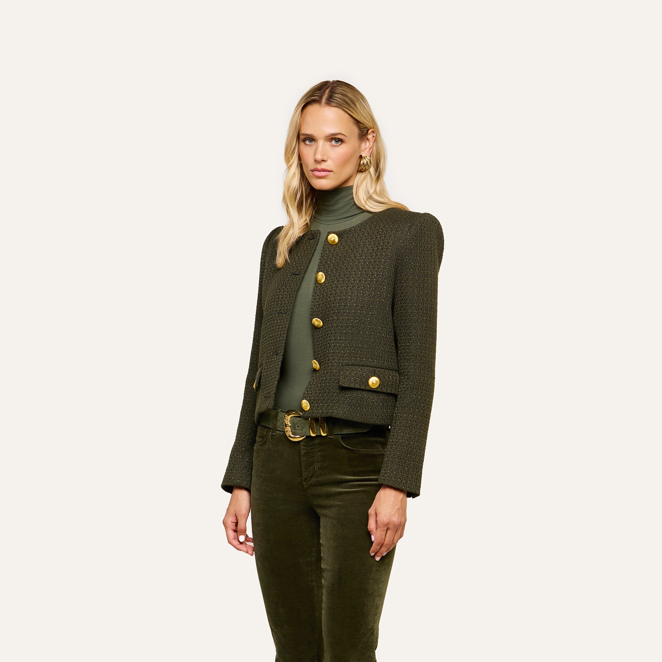 A woman with long blonde hair wears the Zell Tweed Jacket—a dark green style with gold buttons—paired with a matching green turtleneck and green velvet pants, standing against a plain light background.
