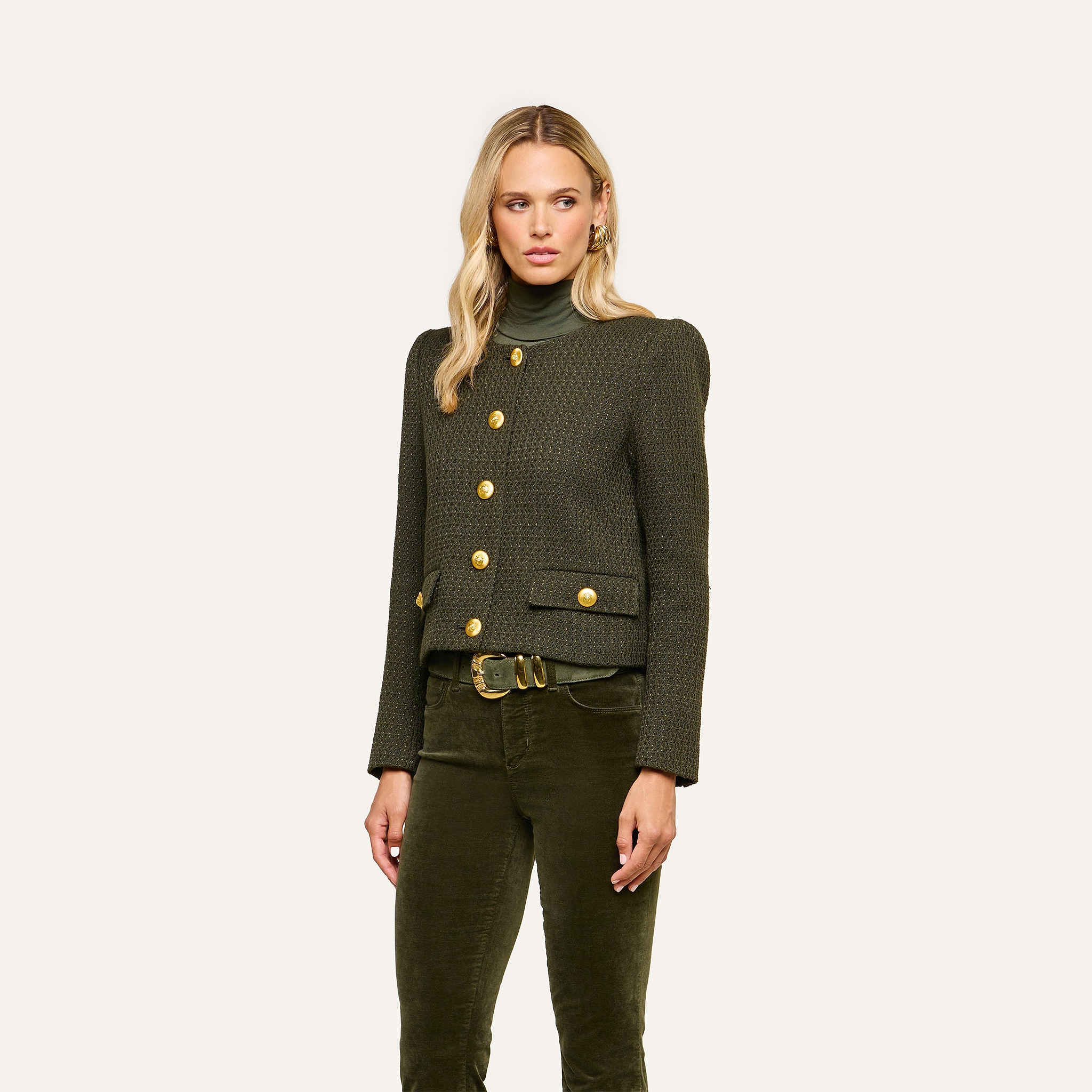 A woman wearing the Zell Tweed Jacket—a dark green, collarless tweed jacket with gold buttons—paired with an olive green turtleneck and matching corduroy pants, stands against a plain light background.
