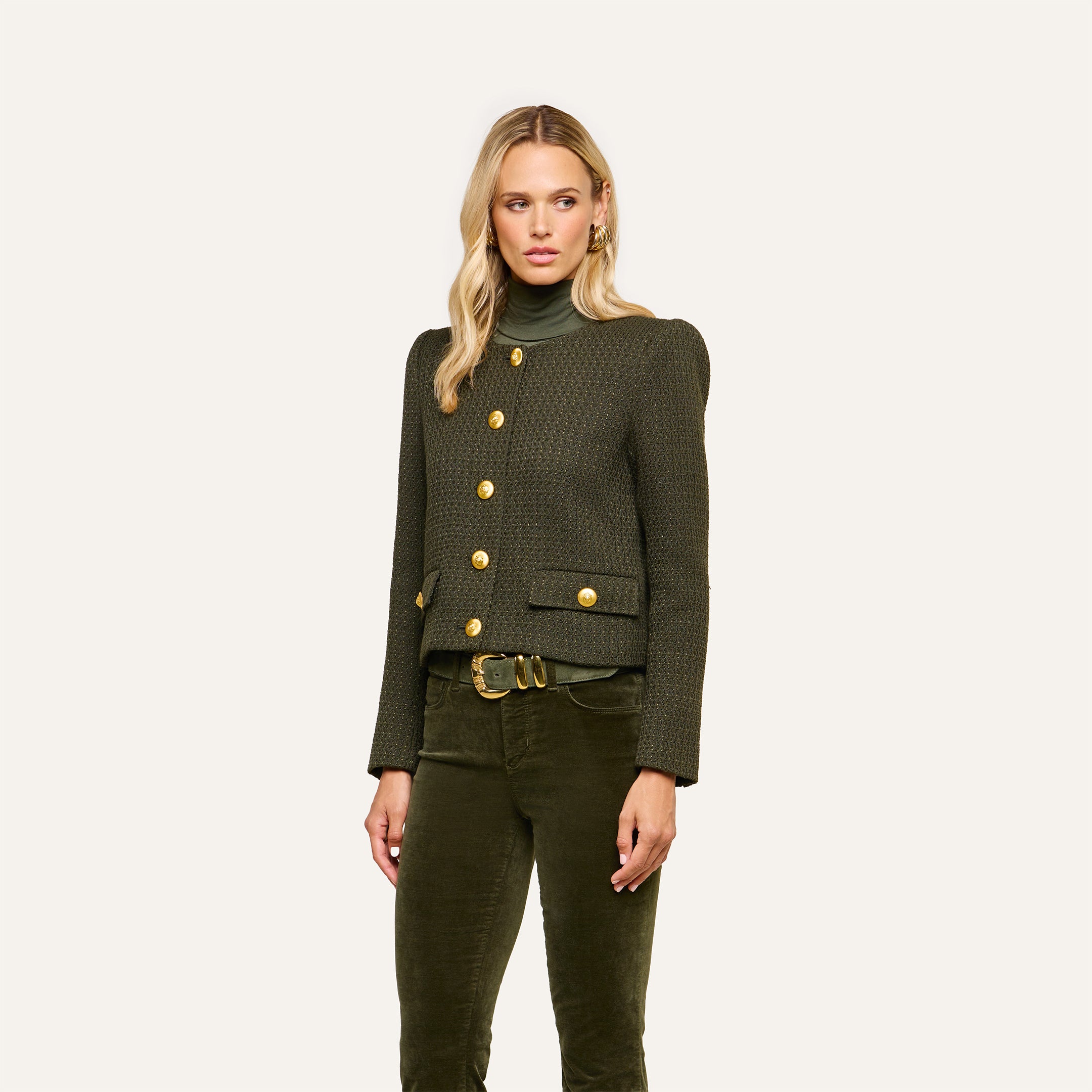 A woman wearing the Zell Tweed Jacket—a dark green, collarless tweed jacket with gold buttons—paired with an olive green turtleneck and matching corduroy pants, stands against a plain light background.