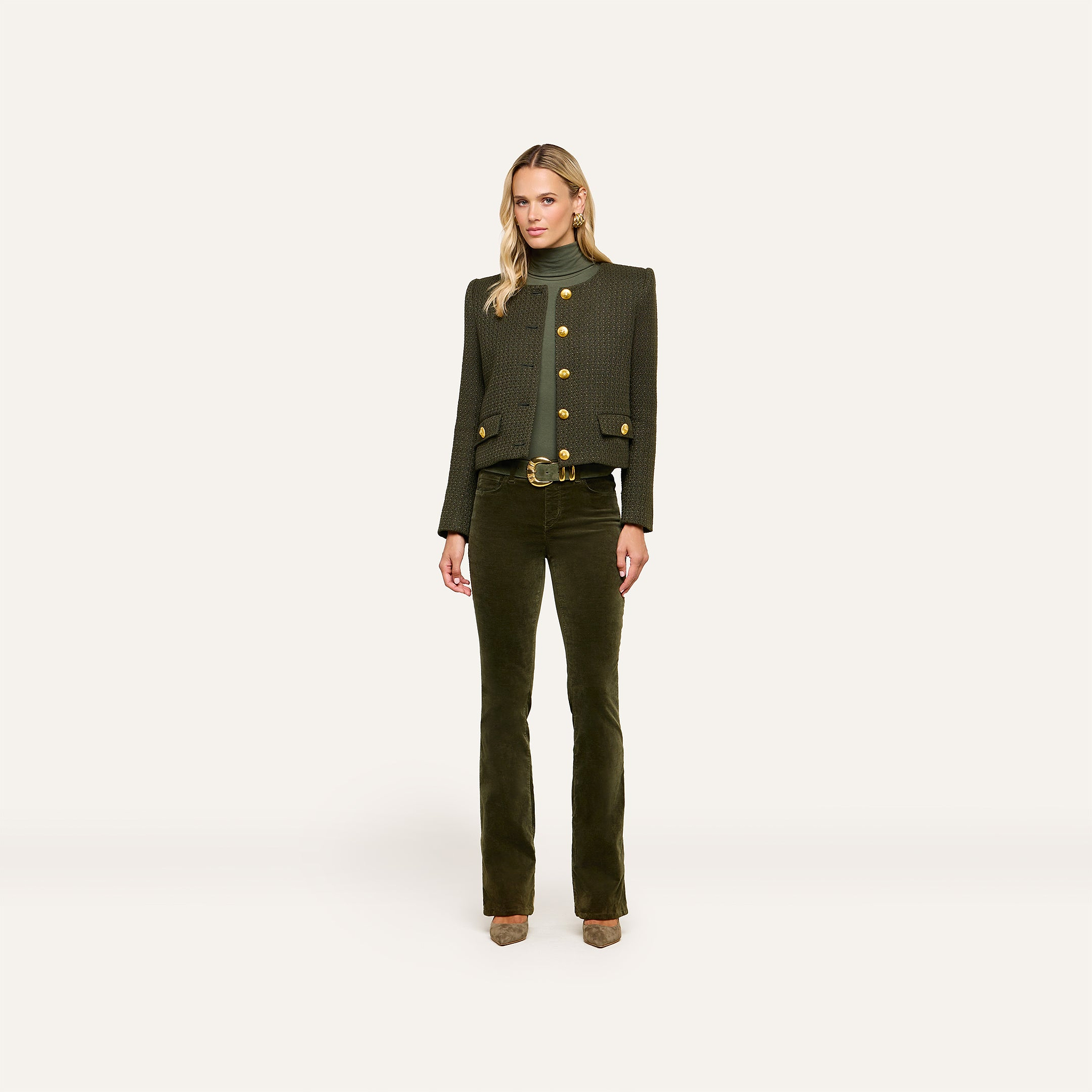A woman stands against a plain background, wearing the dark green Zell Tweed Jacket with gold buttons, matching high-waisted pants, a green turtleneck, and tan heels.