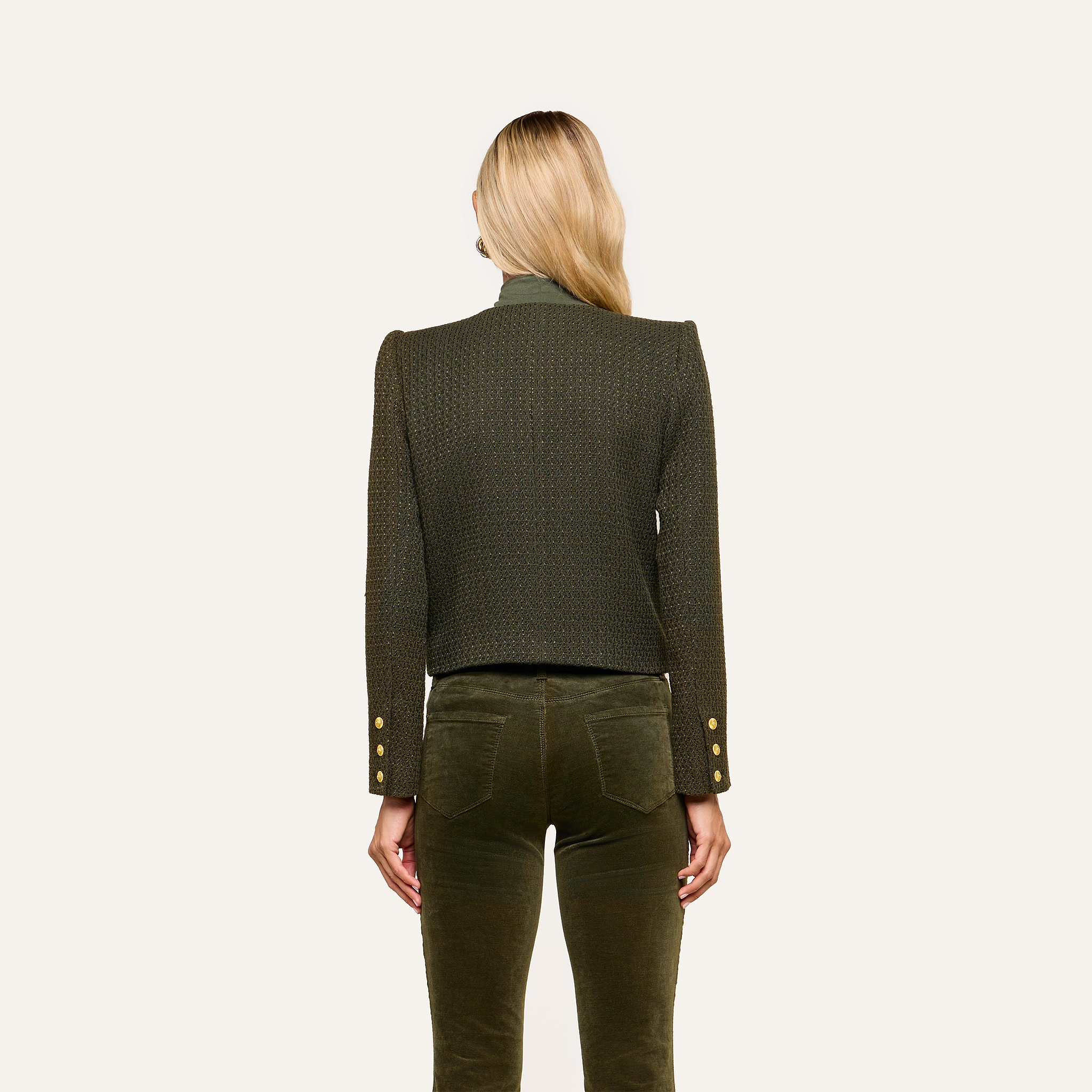 A woman with long blonde hair stands facing away, wearing the Zell Tweed Jacket—a textured, dark green tweed piece featuring gold buttons on the sleeves—paired with matching green pants. The background is plain and light-colored.