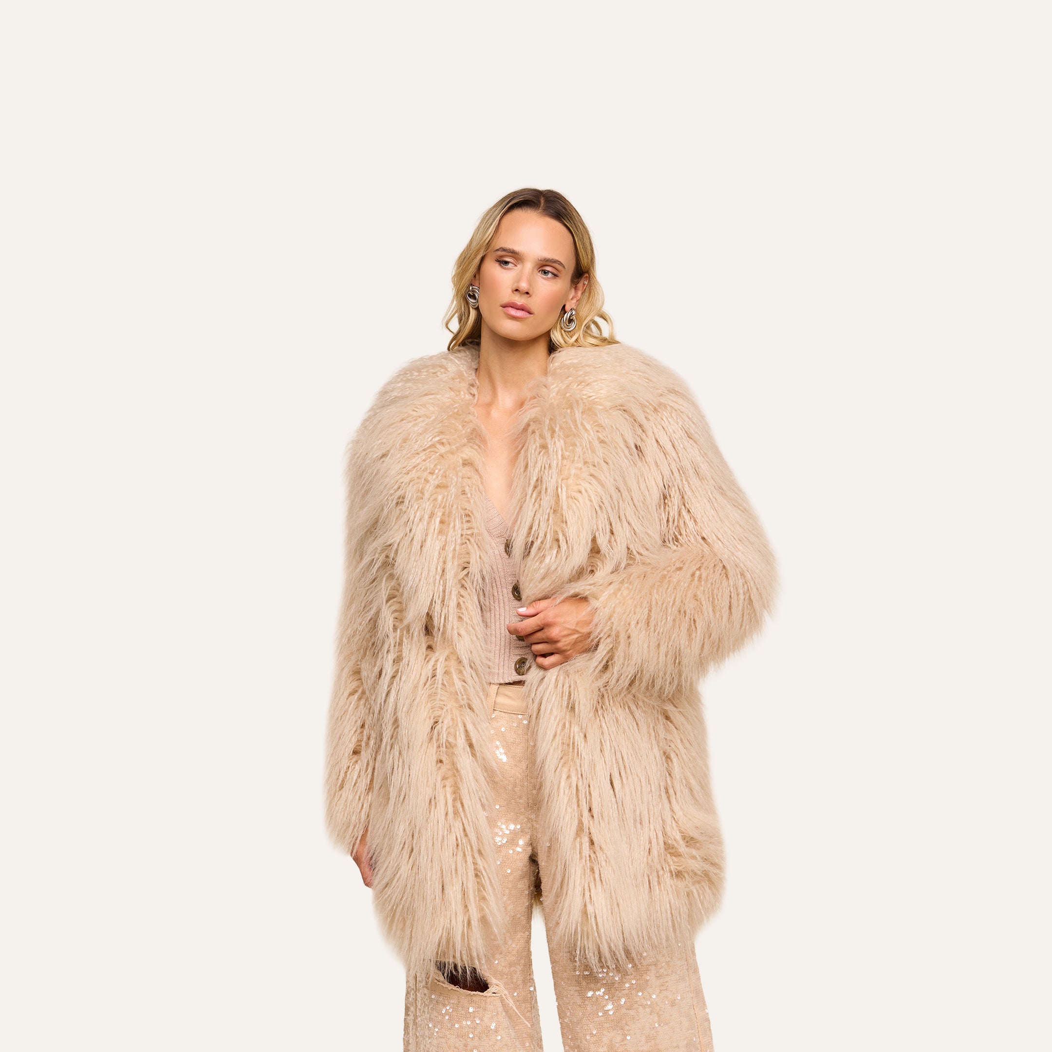 Wearing the Toira Faux Fur Coat over a coordinated beige ensemble with sequin-embellished pants, this individual stands against a plain, light background, effortlessly channeling a vintage-inspired 70s rockstar vibe.