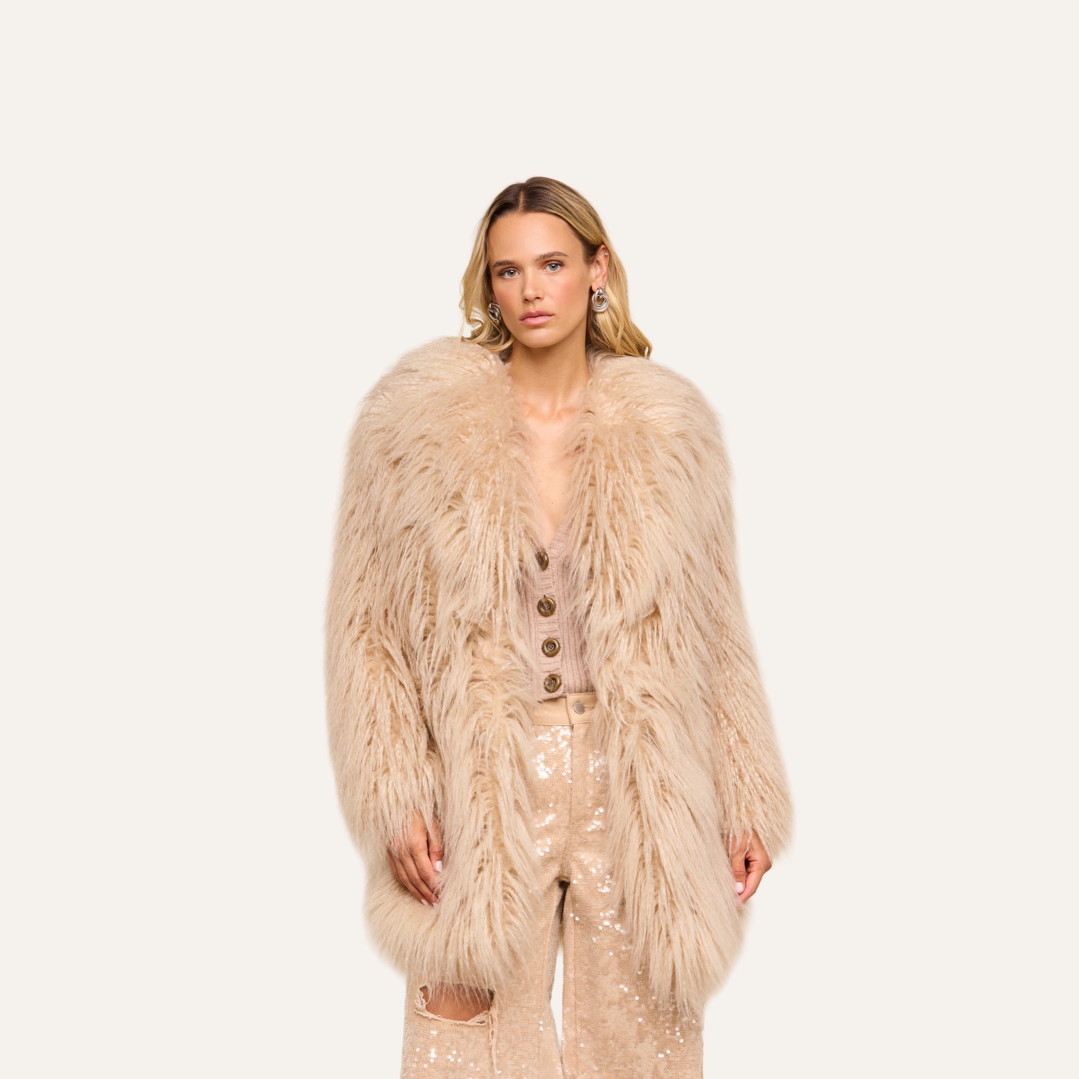 A woman stands facing forward, wearing the Toira Faux Fur Coat—a long, light beige piece—layered over a beige buttoned top and shiny beige sequin pants, channeling a vintage-inspired 70s rockstar vibe against a plain light background.