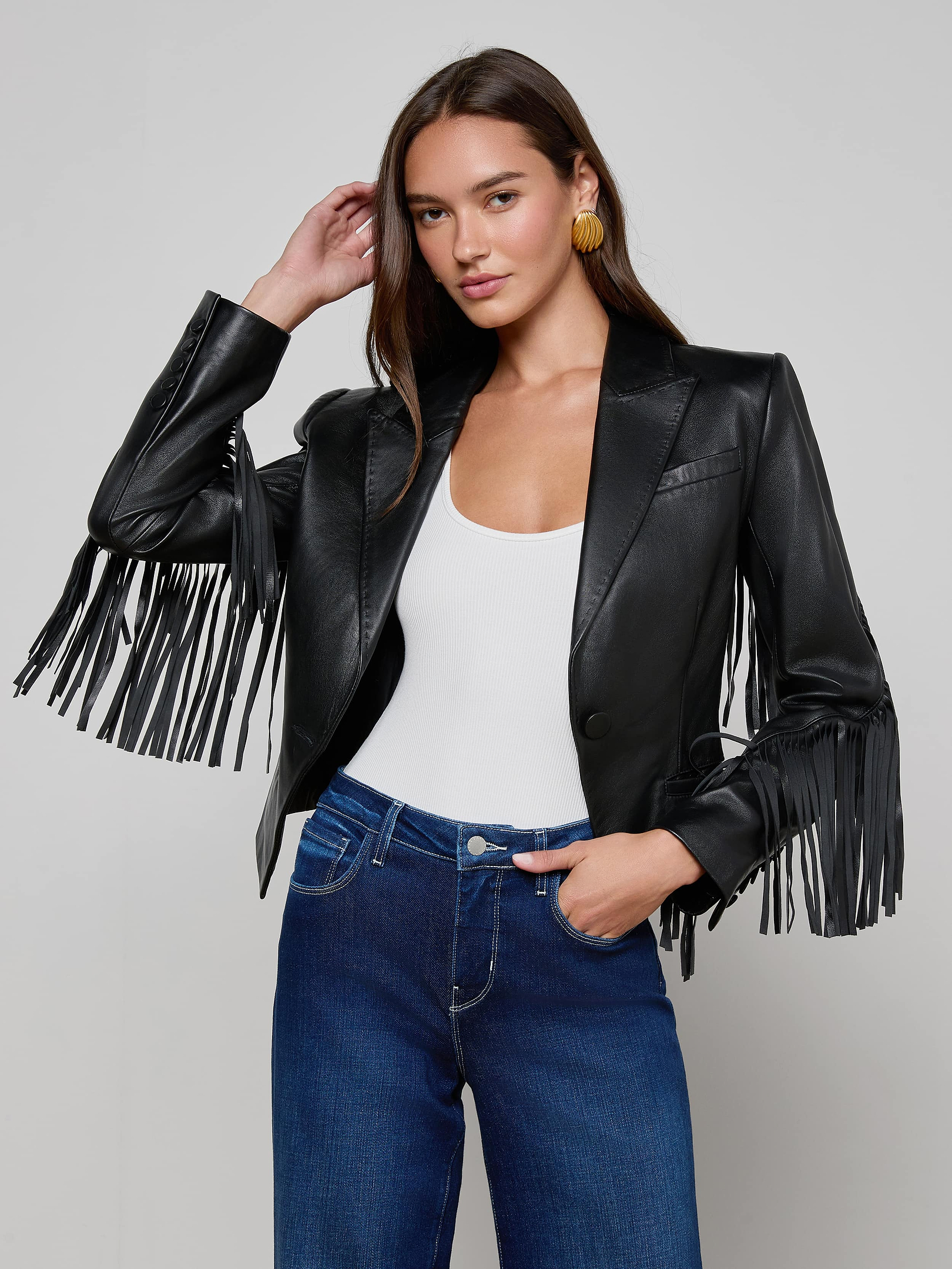 A woman wearing the Vashti Fringed Leather Blazer, styled with a white top and blue jeans, stands against a plain light background.