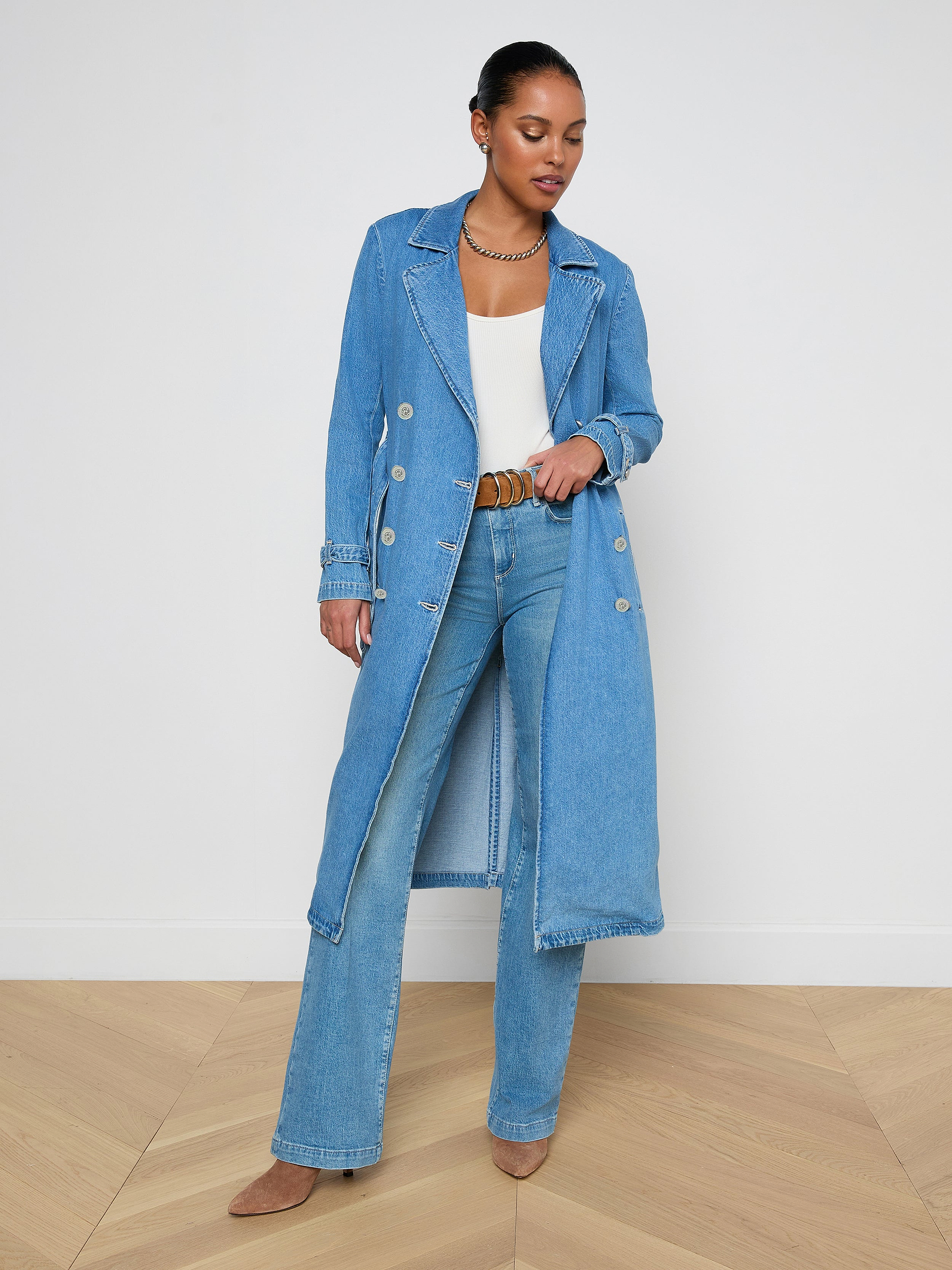 A woman stands wearing the Bradley Trench Coat in a lightweight, medium wash denim over a white top and blue jeans with brown shoes, looking down and gently holding her coat open.