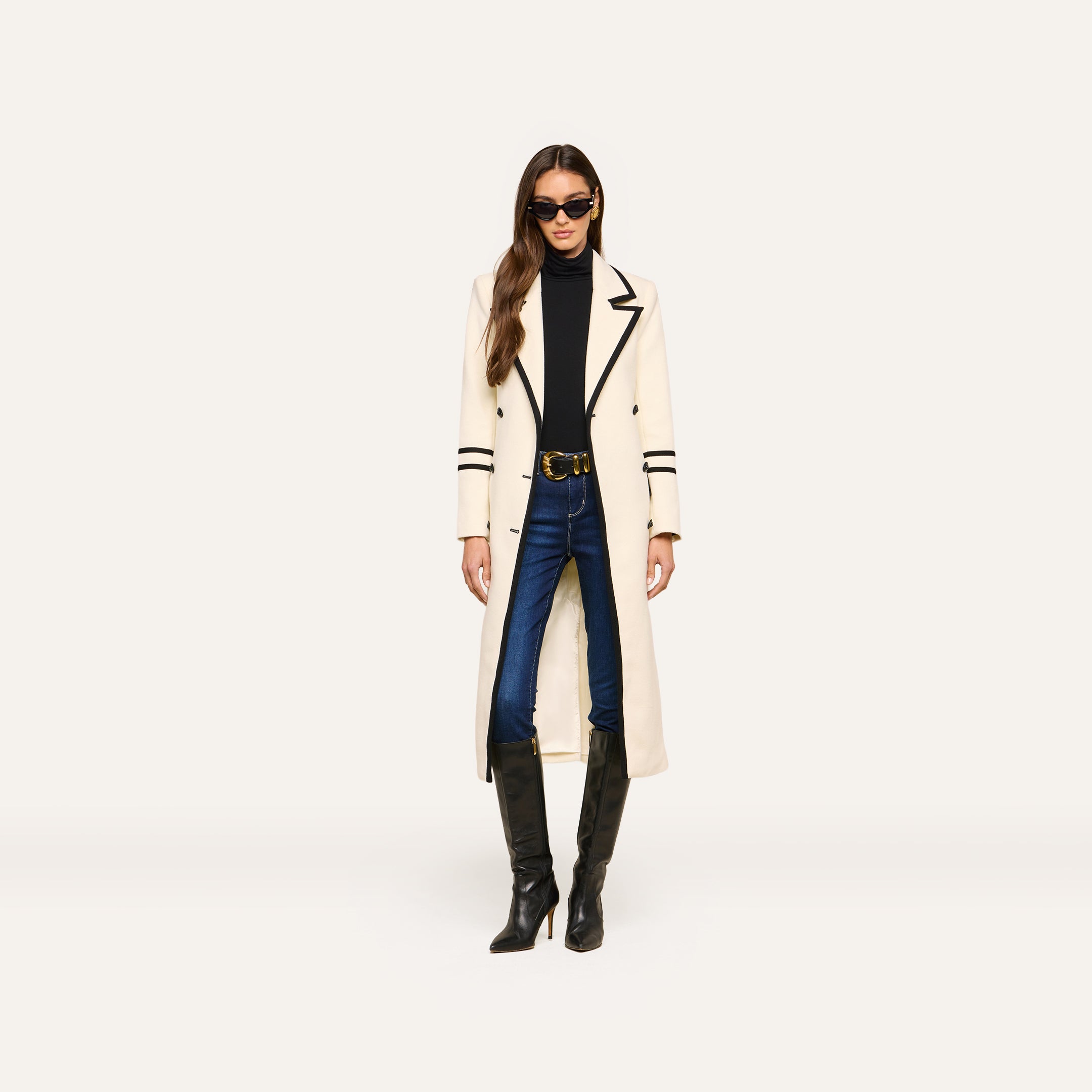 A woman wearing sunglasses, the Irene Long Coat in ivory with black trim, a black turtleneck, blue jeans, and tall black boots stands against a plain white background.