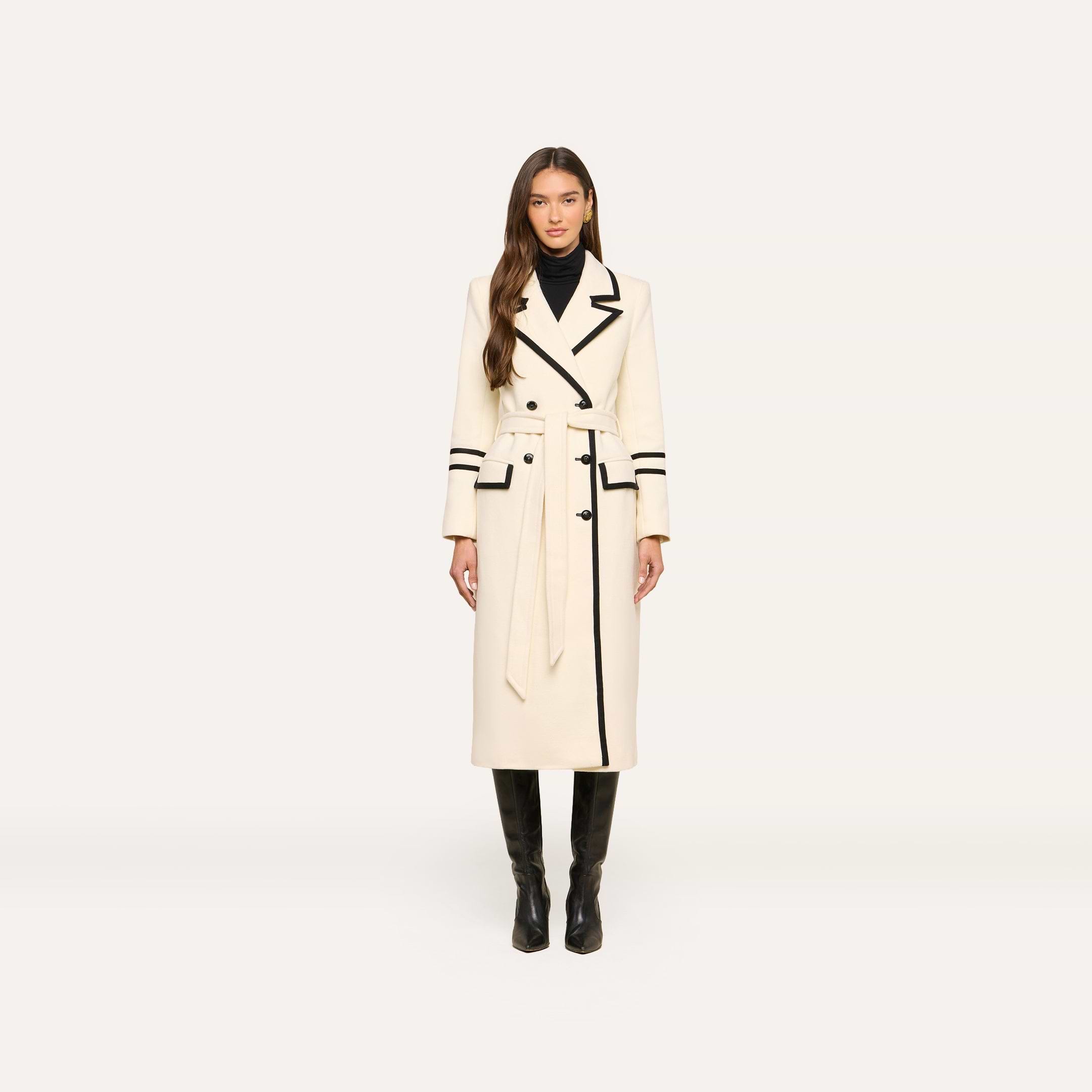A woman stands facing forward against a plain light background, wearing the Irene Long Coat—a double-breasted long ivory coat with black trim—paired with a black turtleneck and knee-high black boots.