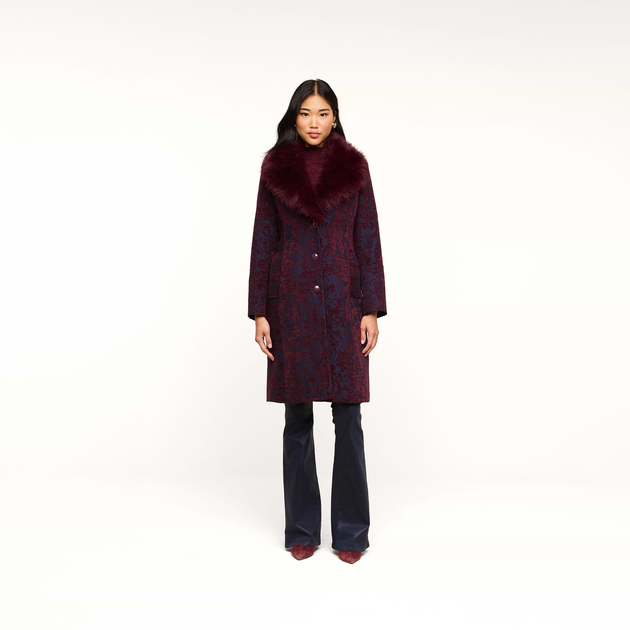 A woman stands facing forward against a plain white background, wearing the Laney Jacquard Coat featuring a vintage-inspired design with a large burgundy faux fur collar, paired with navy pants and burgundy shoes.