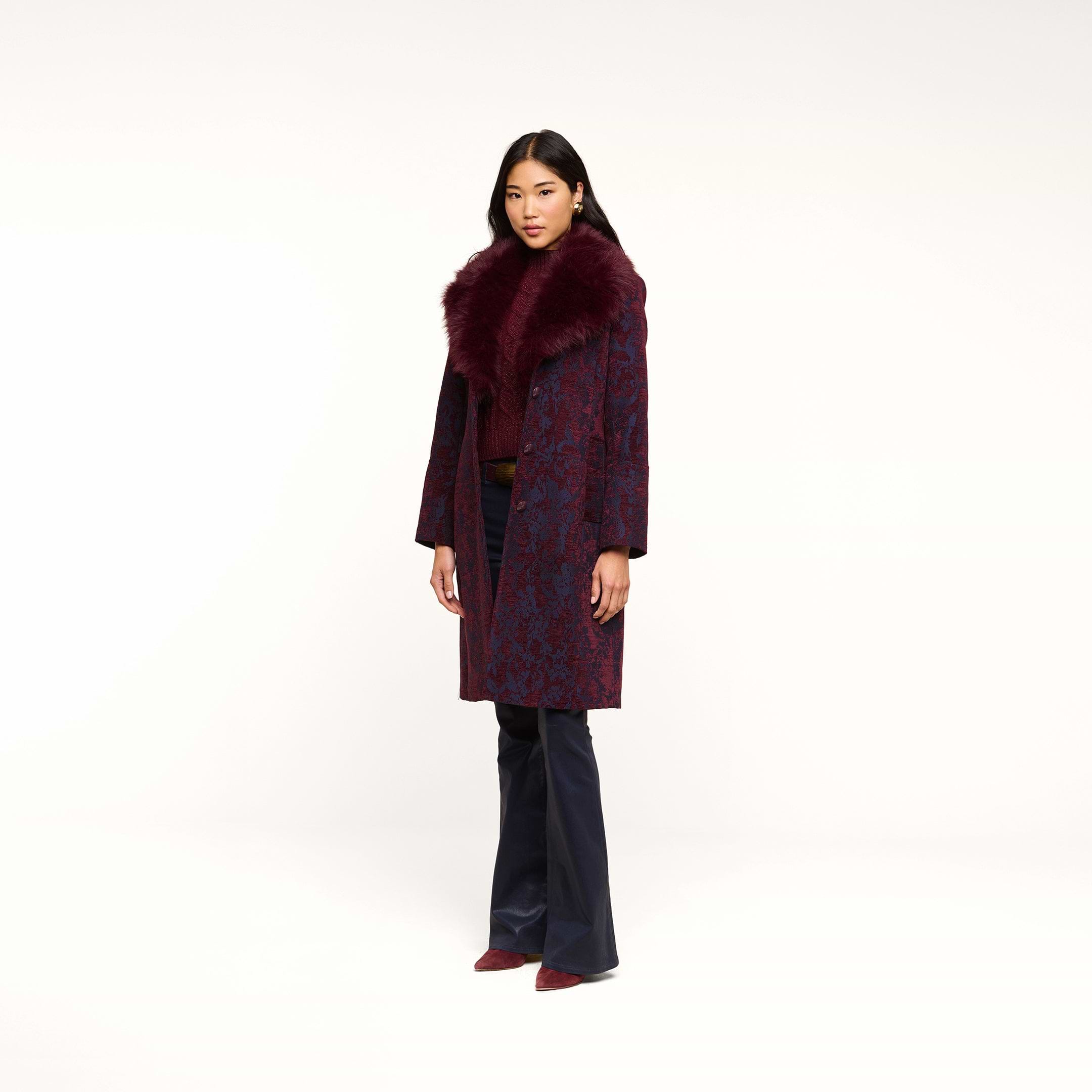 A woman stands against a plain white background, wearing the Laney Jacquard Coat—a long, vintage-inspired coat featuring a large faux fur collar—paired with dark pants and burgundy shoes.