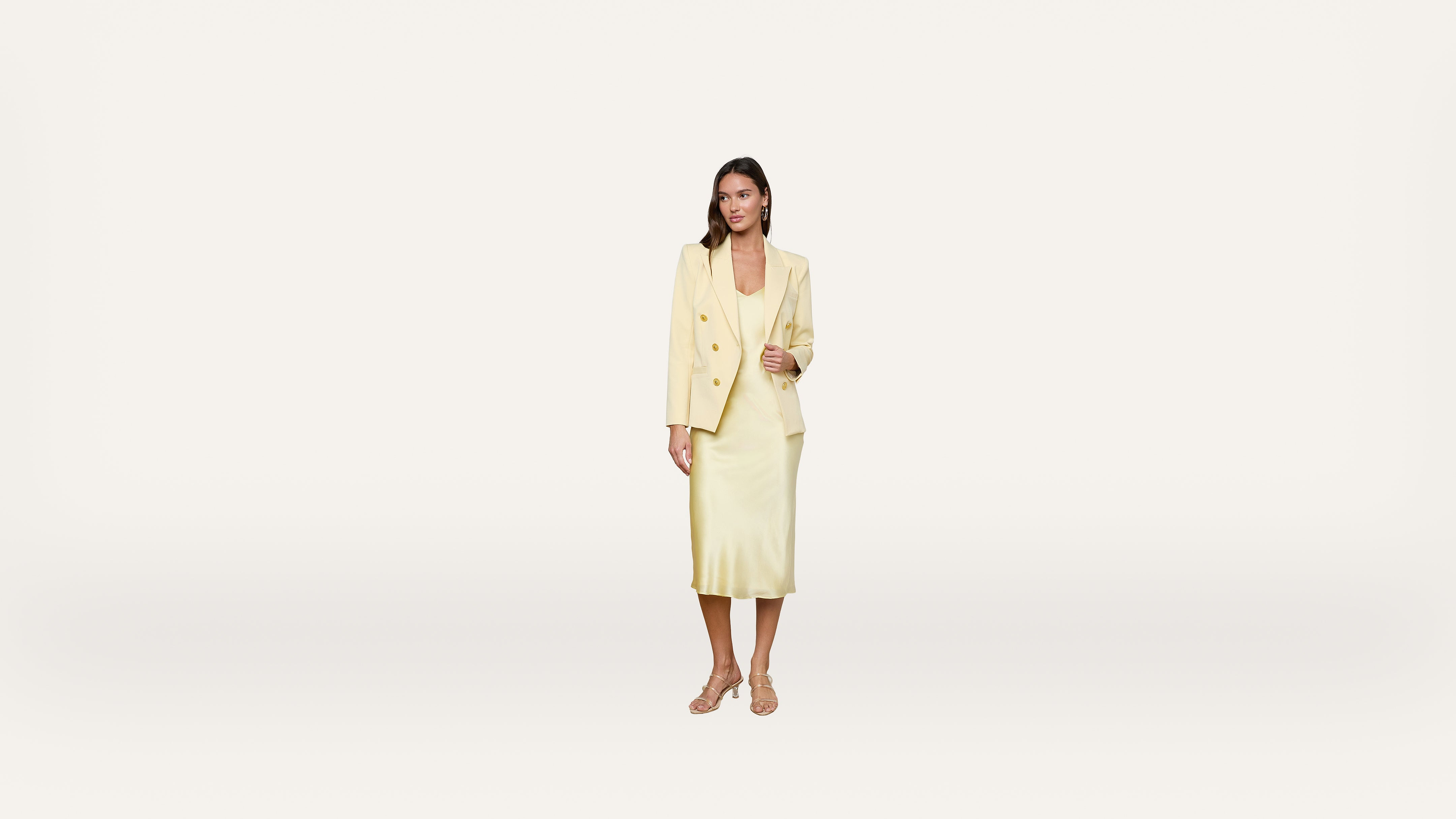 A woman stands against a plain white background wearing a light yellow blazer and matching midi dress with strappy sandals.
