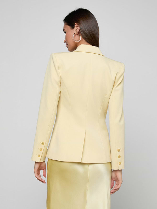 A woman is shown from the back wearing the Kyomi Double-Breasted Blazer in yellow with gold buttons and a matching satin skirt, standing against a plain off-white background.