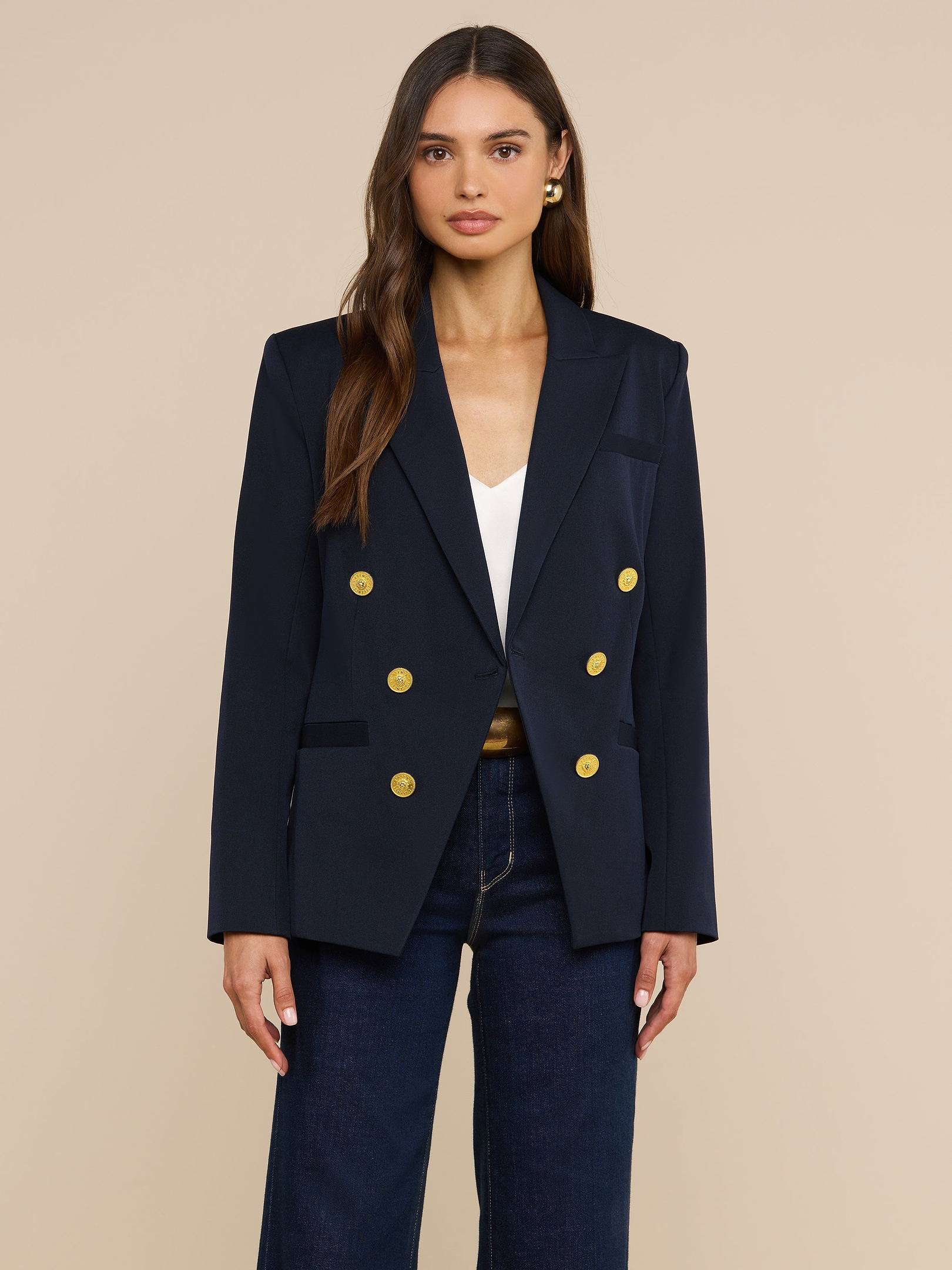 A woman with long brown hair wears the Kyomi Double-Breasted Blazer in midnight navy, featuring gold buttons, over a white top and dark jeans, standing against a plain light background.