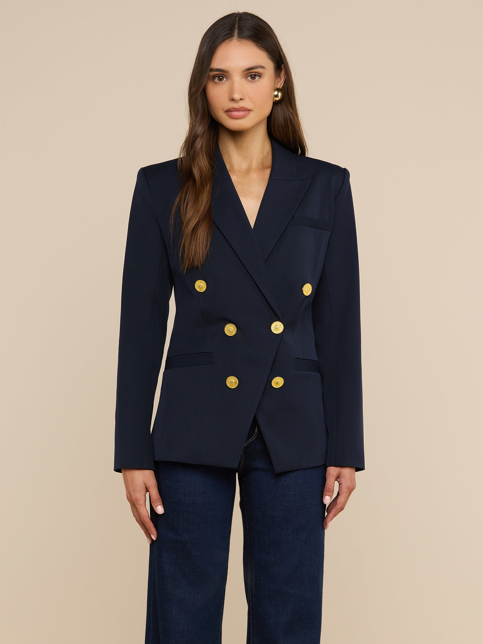A woman stands facing forward, wearing the Kyomi Double-Breasted Blazer in midnight navy with gold buttons and dark pants, against a plain light background.
