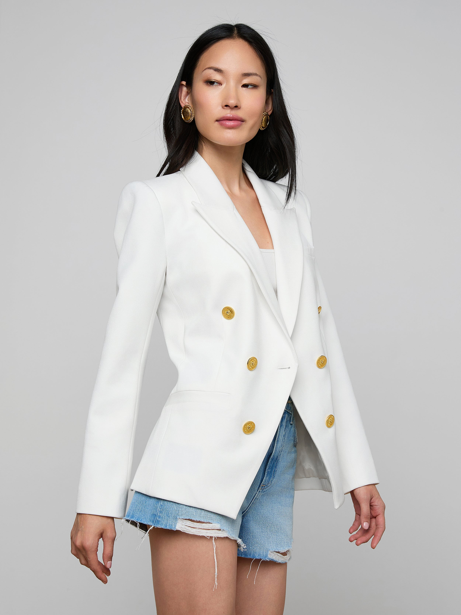 A woman wearing the Kyomi Double-Breasted Blazer in white, crafted from stretch suit fabric, paired with distressed denim shorts, stands against a plain light background.