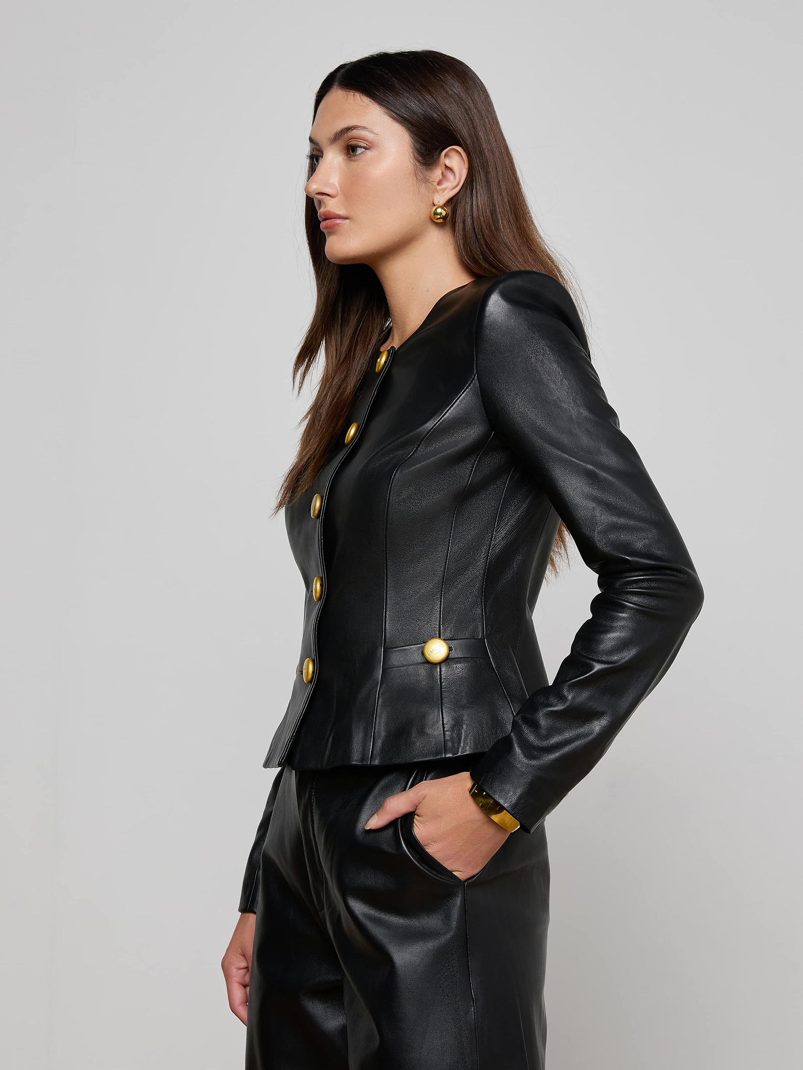 A woman in the Mariella Leather Jacket—a fitted, collarless lambskin piece with gold buttons—paired with matching pants stands sideways against a plain light background. She has straight brown hair and accessorizes with gold earrings and a bracelet.